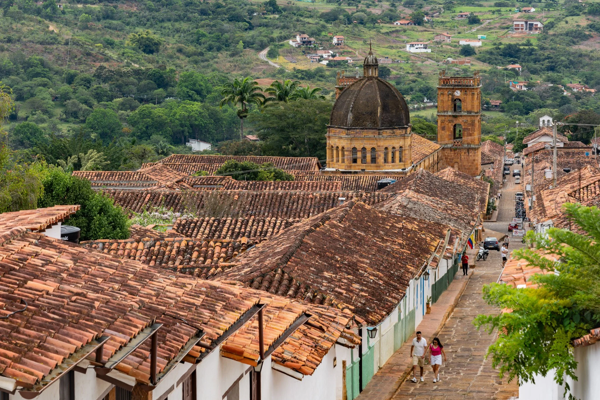 Vista de una ciudad colonial con techos de tejas rojas y una iglesia con cúpula central y torre del reloj, rodeada de vegetación y montañas de fondo, con personas caminando por la calle adoquinada.