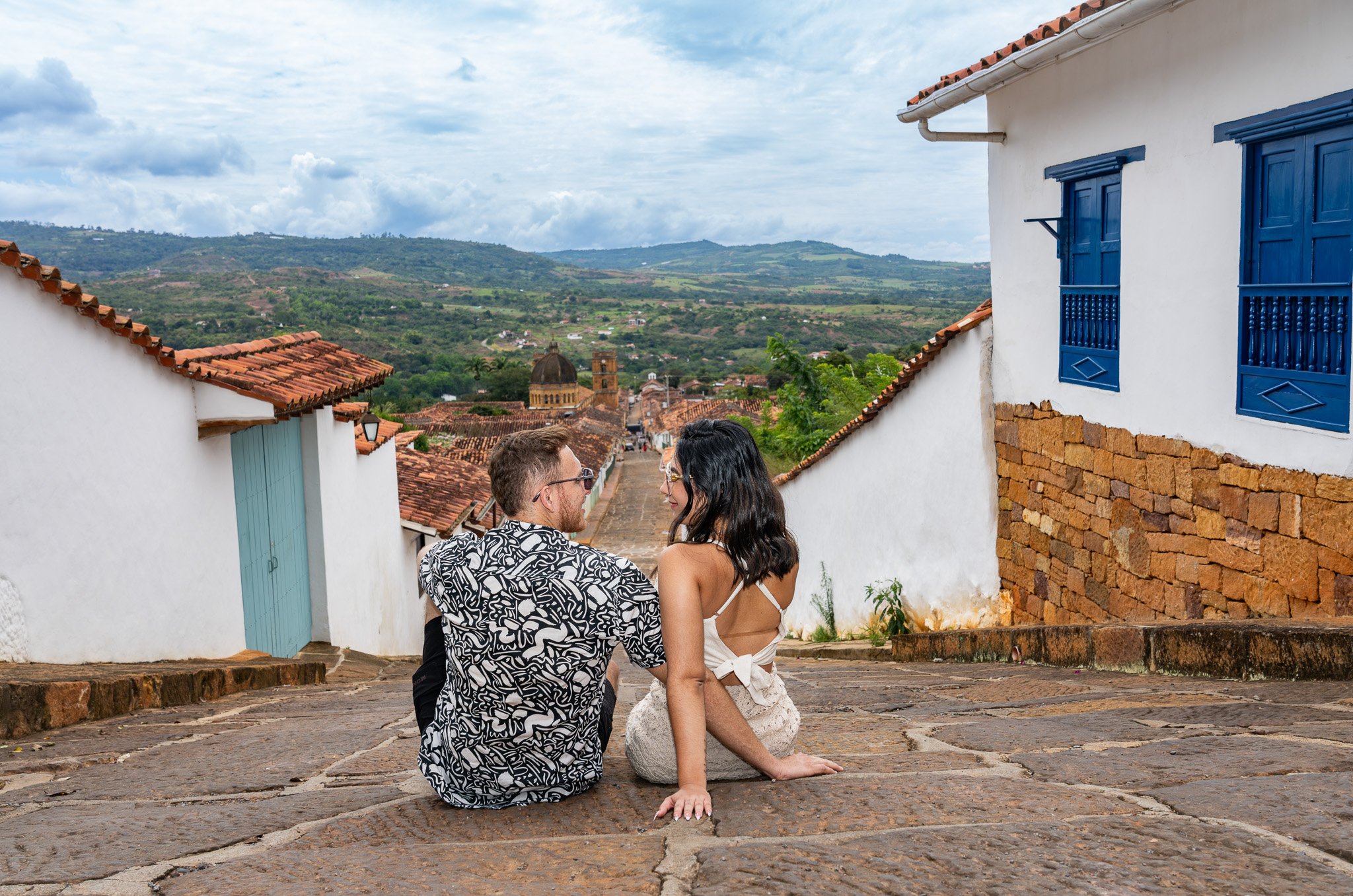 Pareja sentada en las calles de una localidad con casas blancas y ventanas azules, vistas verdes y montañas al fondo.