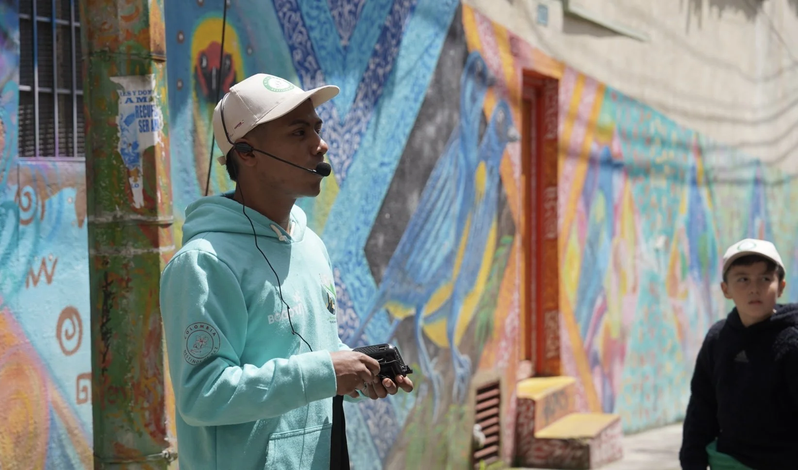 Joven con auriculares y casco, sosteniendo un dispositivo, frente a un mural colorido en la pared de un edificio, con otro joven de fondo.