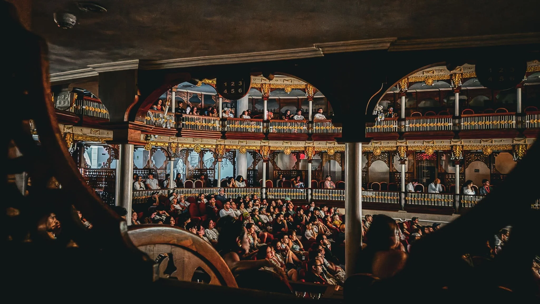 Audiencia en un teatro con múltiples niveles de balcones decorados con detalles dorados y madera, llena de personas sentadas mirando hacia adelante.