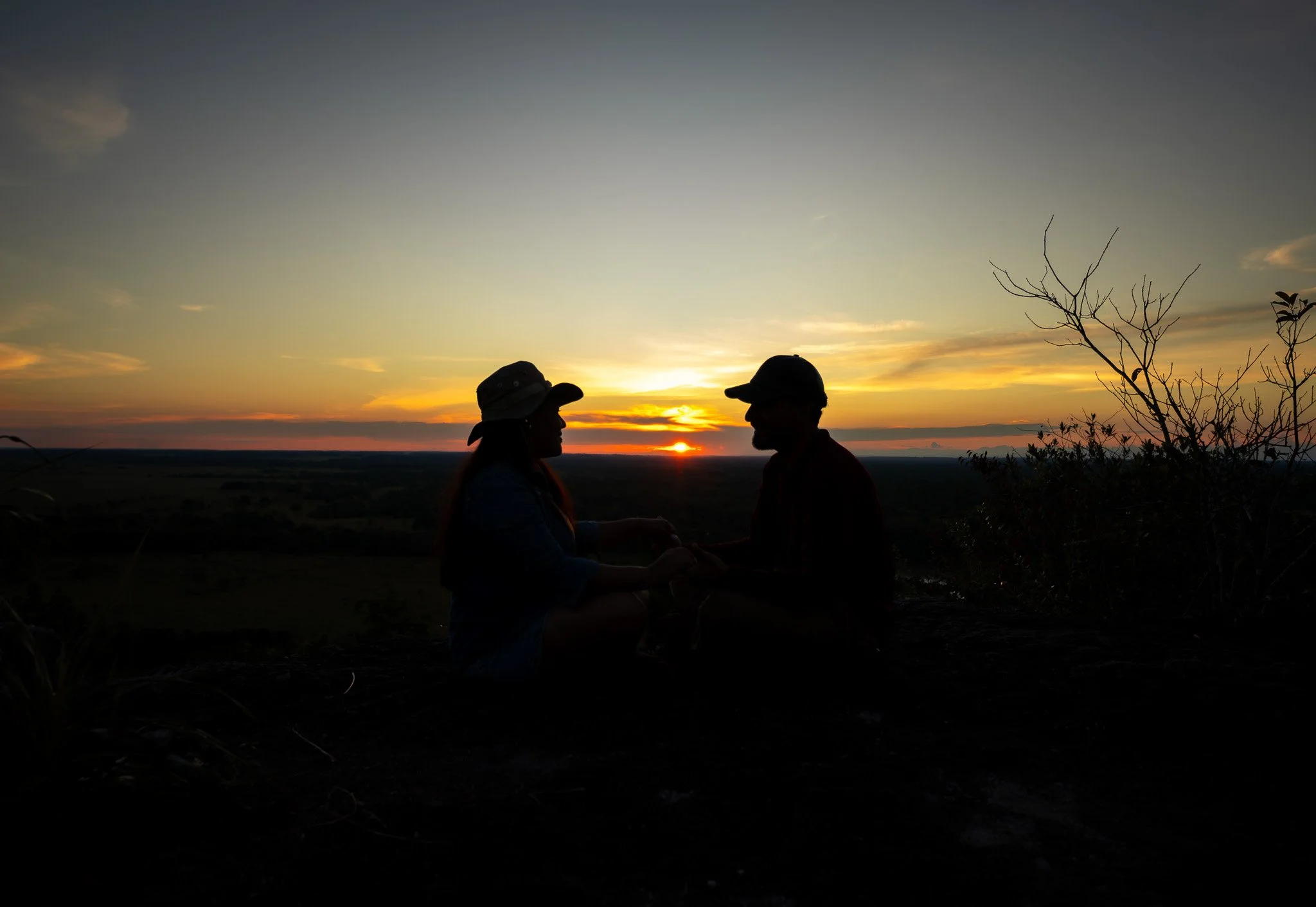 Dos personas con sombreros en silueta, conversando durante un atardecer en un paisaje abierto con cielo despejado y árboles en el fondo.