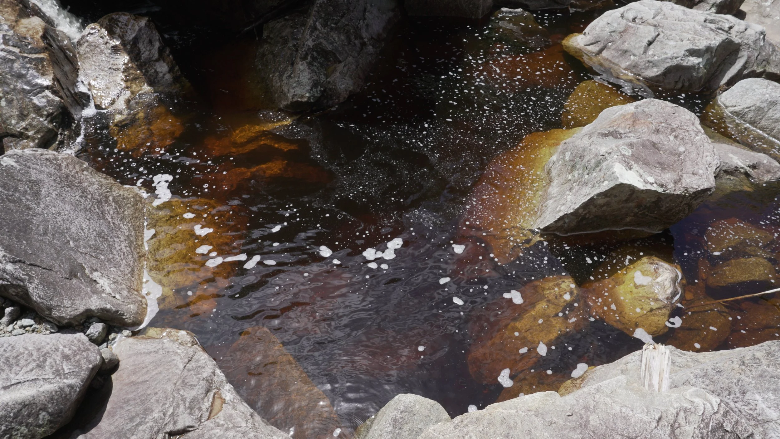 Aguas oscuras en un río rodeado de rocas de diferentes tamaños, algunas con espuma en la superficie.