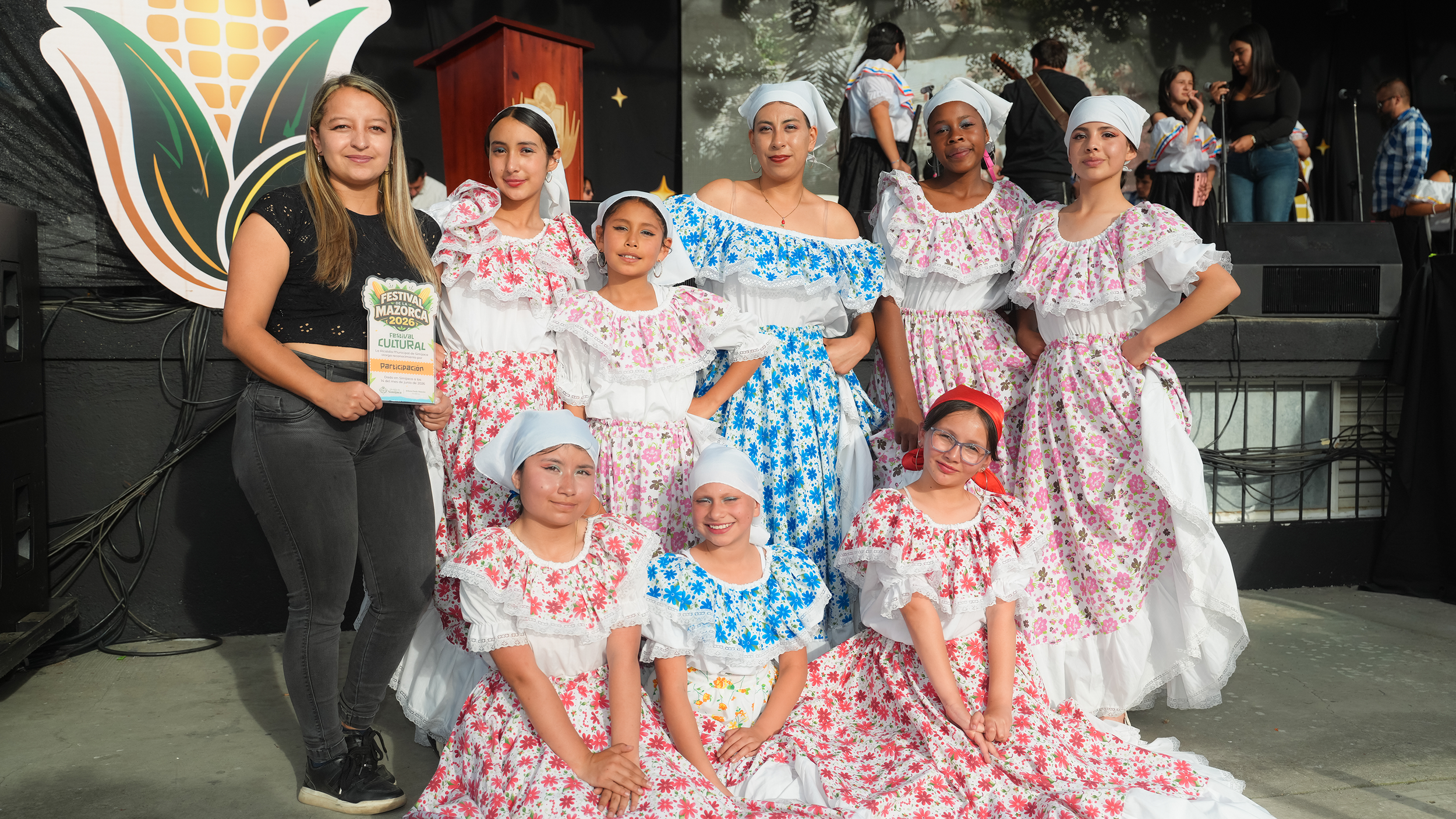 Grupo de personas y niños en un escenario durante un festival cultural, algunos con trajes tradicionales y otras con ropa moderna, en un ambiente festivo.