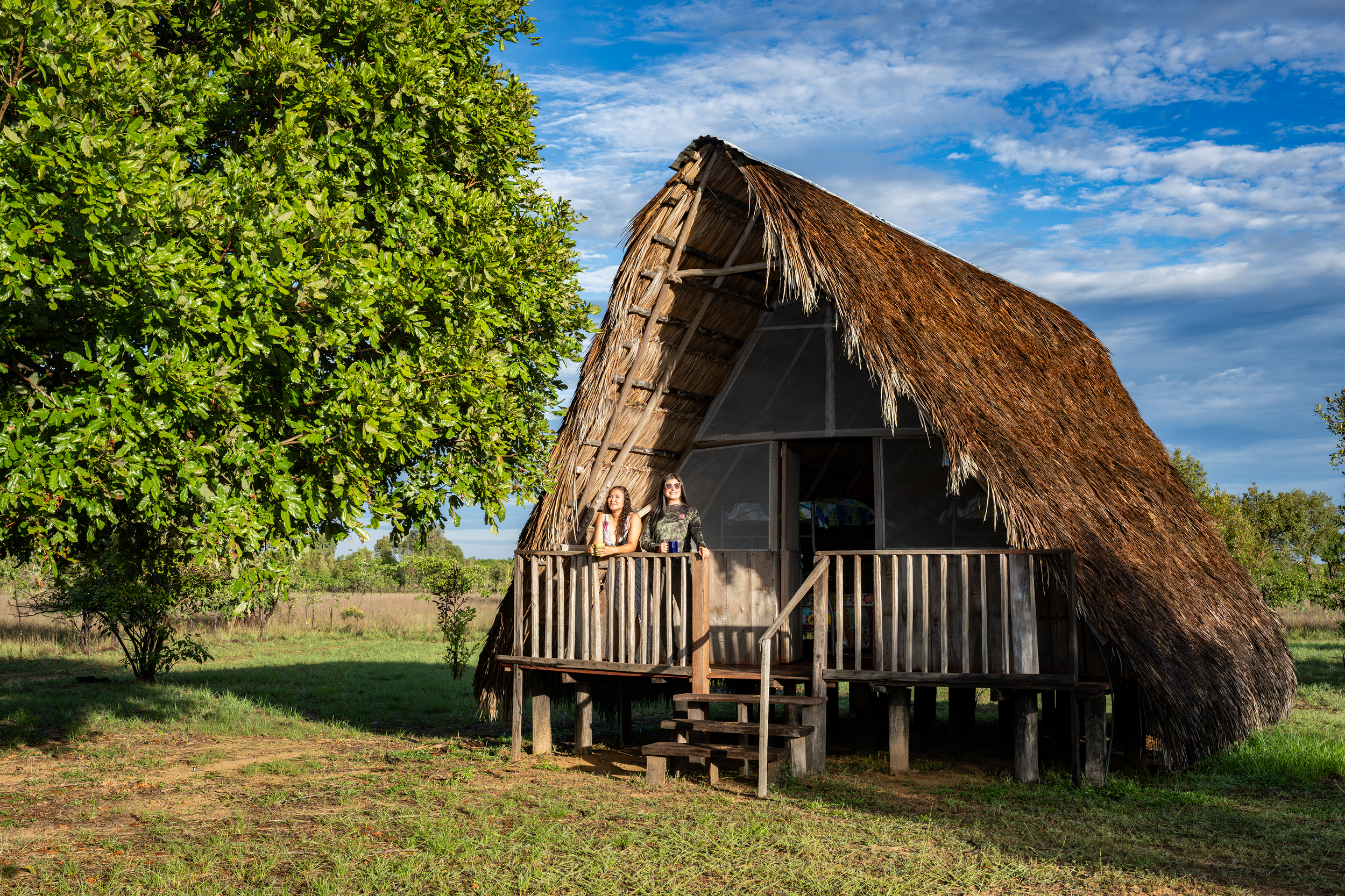 Casita tradicional de madera con techos de paja, ubicada en un campo abierto con árboles verdes y un cielo con nubes, y dos personas en el balcón