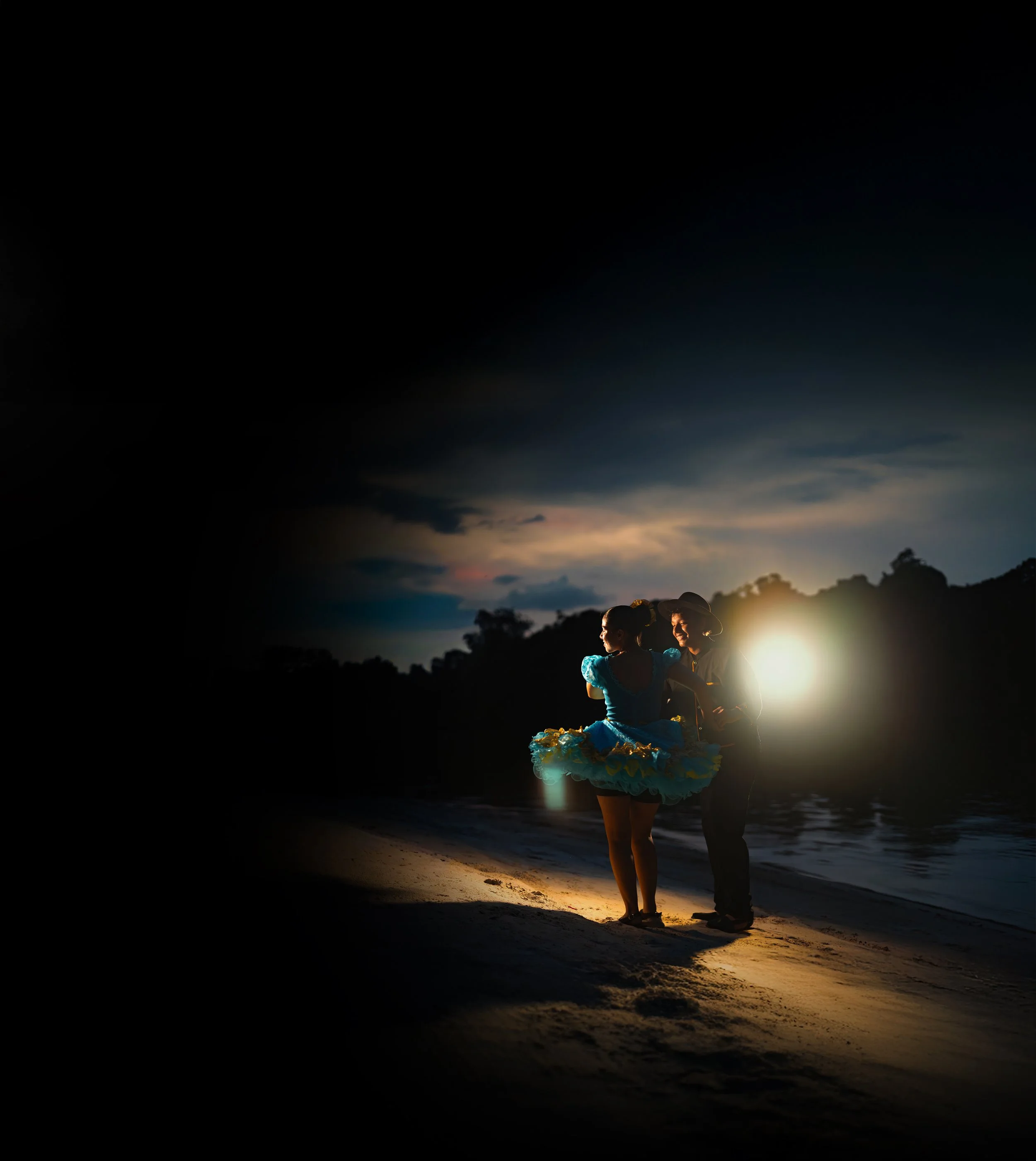 Dos mujeres bailando con vestidos de colores en la playa durante el atardecer, con el sol oculto en el horizonte y árboles en el fondo.