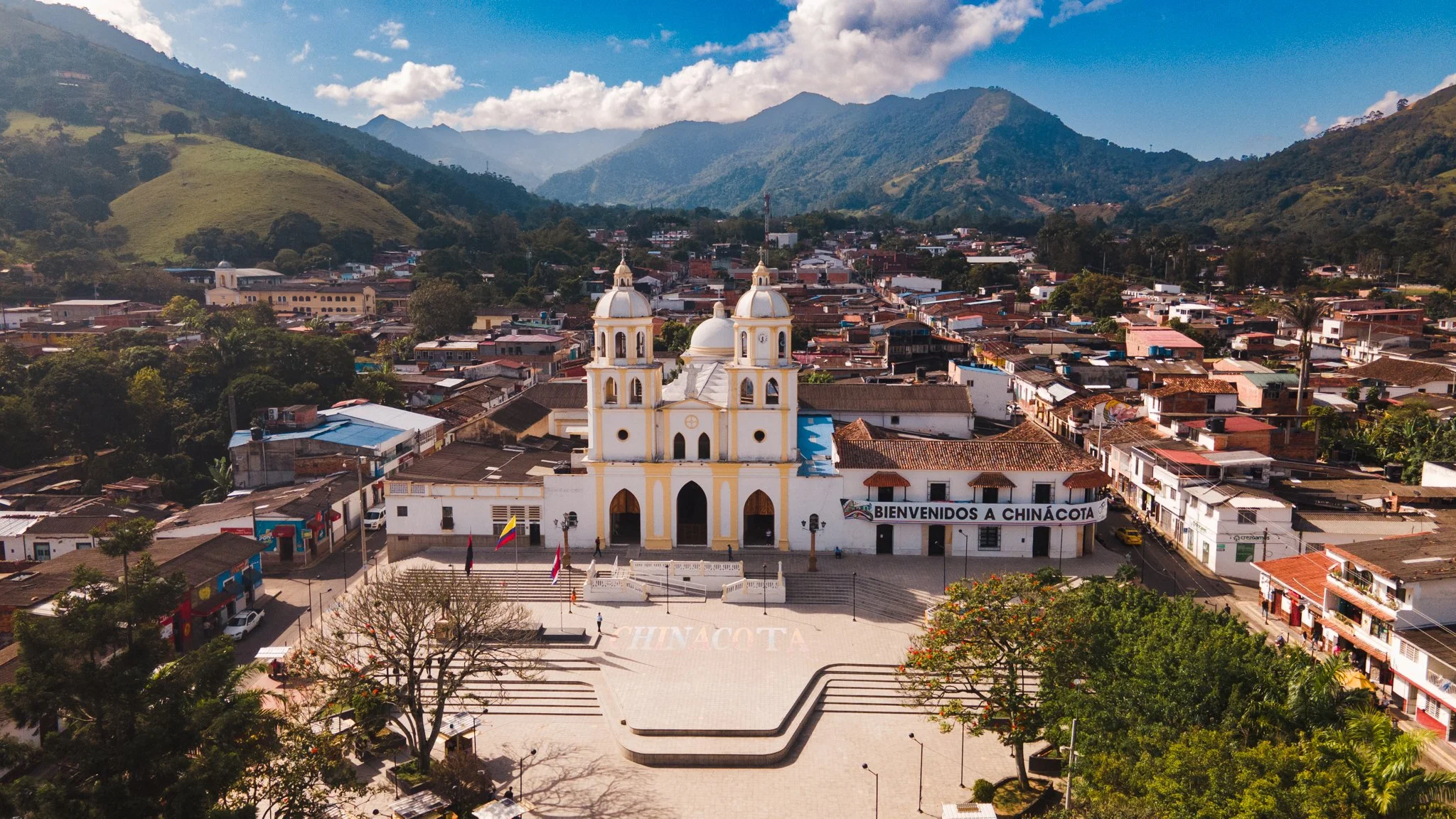 Vista aérea de un pueblo rodeado de montañas, con una iglesia de color amarillo y blanco en primer plano, y un cartel que dice "Bienvenidos a Chinácota" en la zona central.