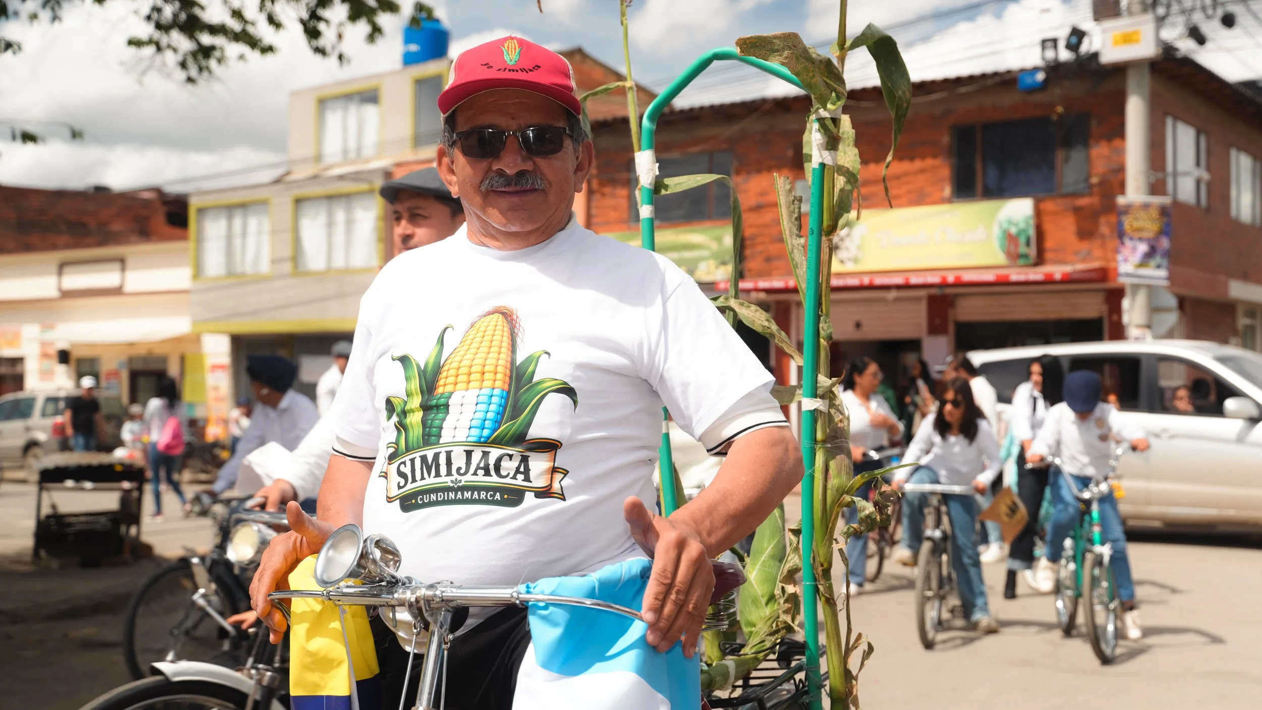 Hombre con gafas y sombrero rojo, paseando en bicicleta durante un evento en una calle con muchas personas y edificios de fondo, lleva camiseta con gráfico de maíz y la leyenda 'SimiJACA Cundinamarca'.