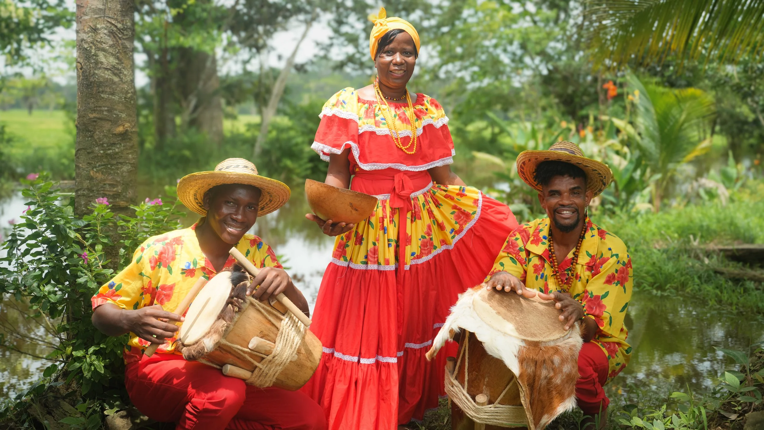 Cuatro personas vestidas con ropa tradicional colorida, tocando instrumentos musicales en un entorno natural con árboles y agua.