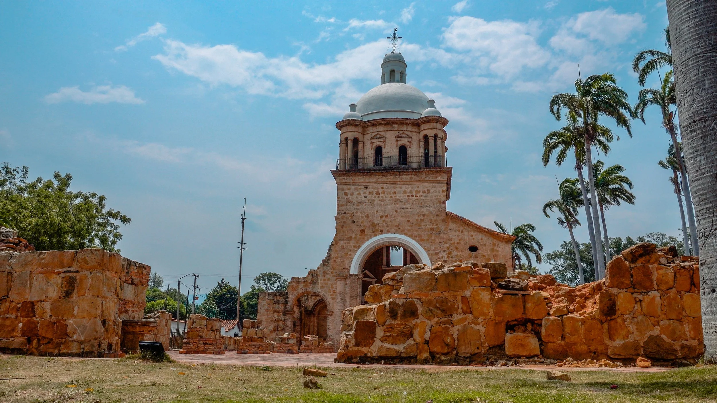 Torre colonial de piedra en ruinas, con muros rotos y restos de ladrillos, rodeada de palmeras y árboles, bajo un cielo azul con nubes.