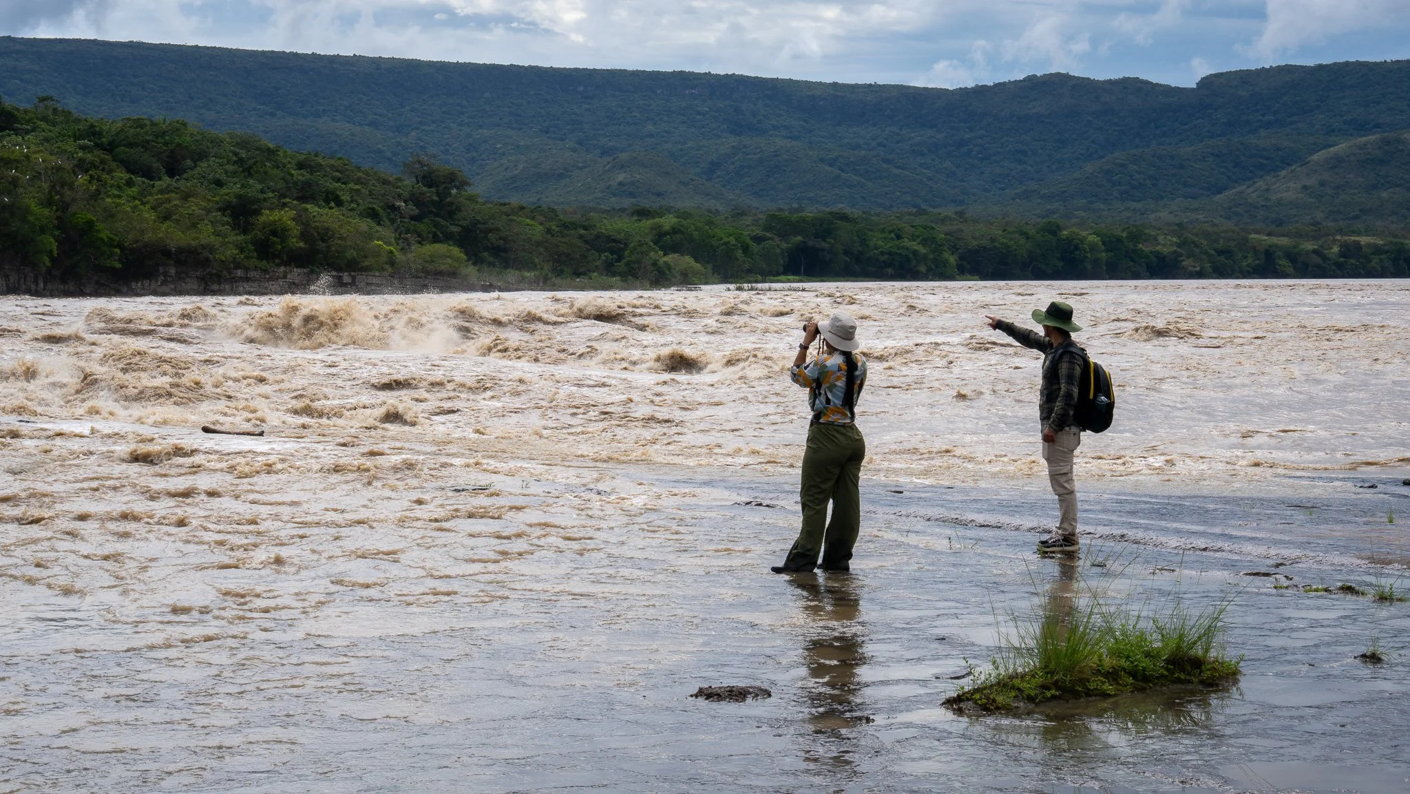 Dos personas de pie en las orillas de un río caudaloso y turbulento rodeado de montañas con vegetación densa. Una de las personas está señalando hacia el río y la otra tomando una foto.