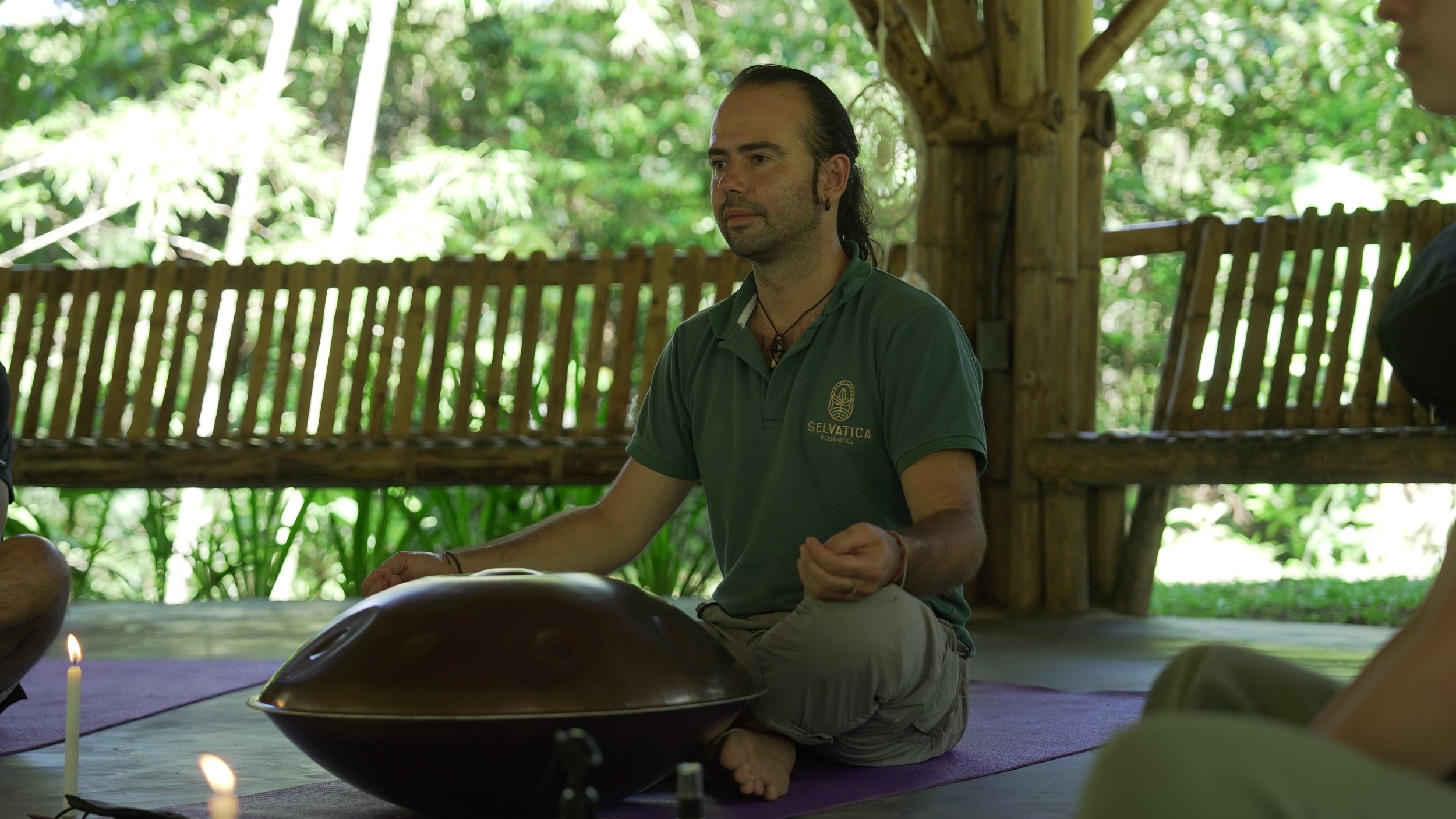 Hombre con vestimenta verde sentado en un ambiente natural con vegetación, tocando un instrumento musical de madera y metal.