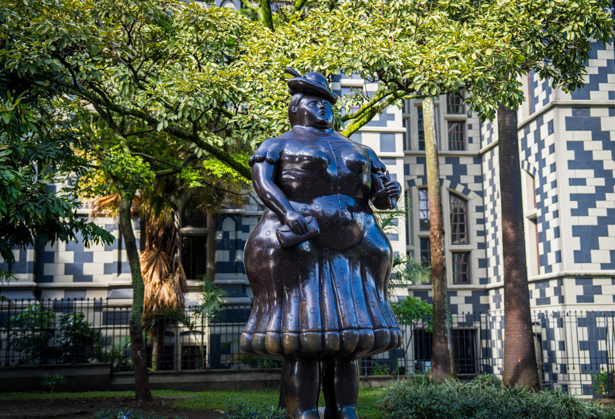 Escultura negra de una mujer con sombrero, en un parque con árboles y en un fondo de edificio moderno con patrones de ladrillos blancos y negros.