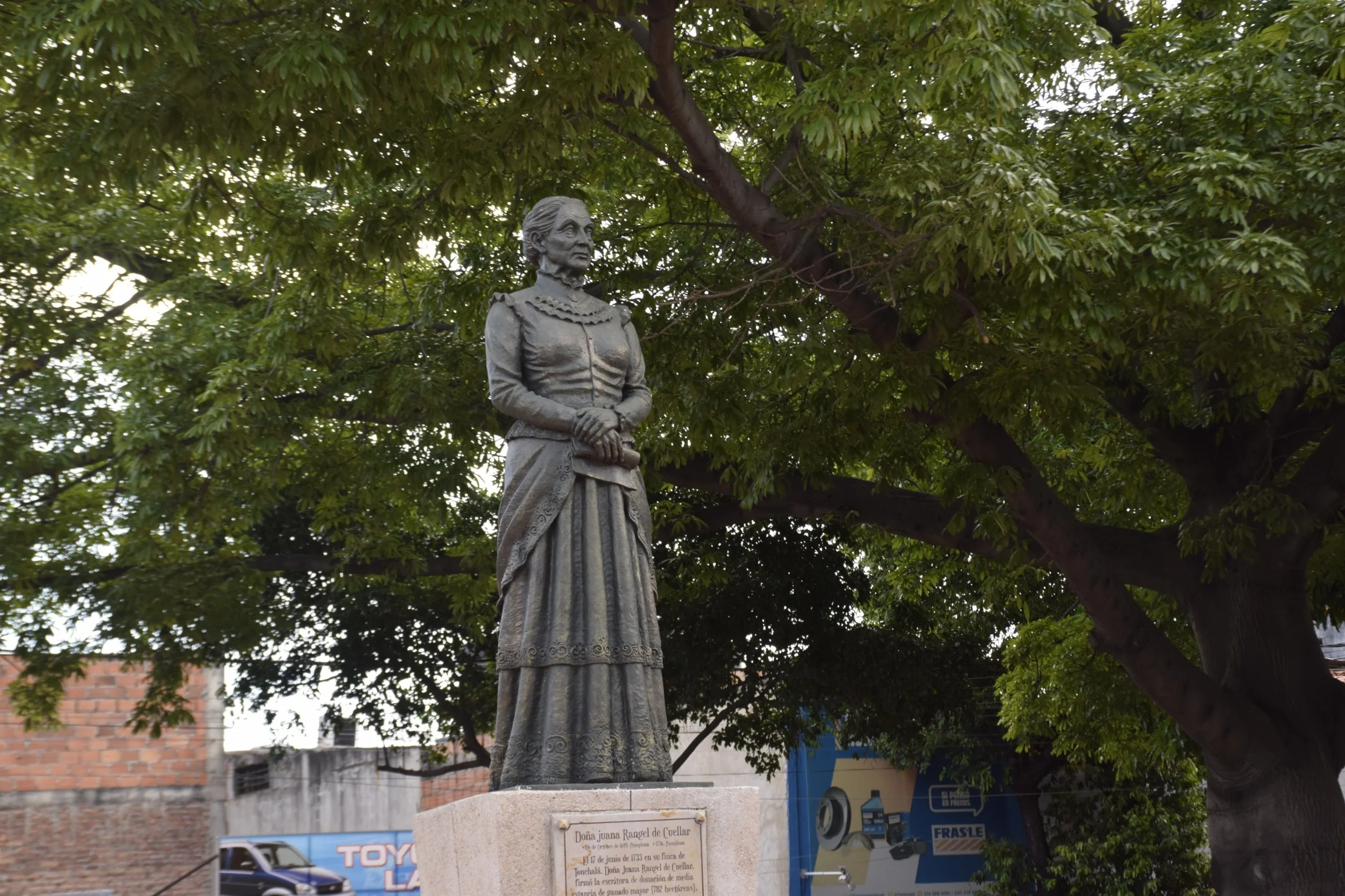 Una estatua de una mujer vestida con ropa tradicional, con cabello recogido, de pie bajo un árbol con muchas hojas verdes, en un entorno urbano con edificios de ladrillo y anuncios publicitarios al fondo.