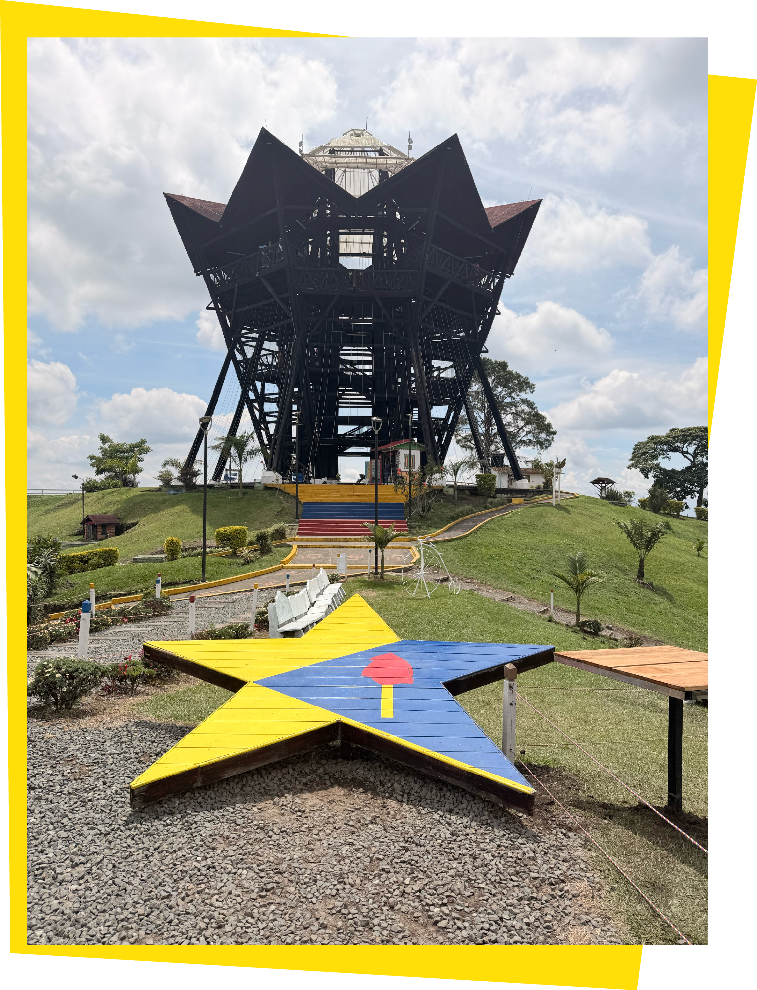 Estructura de madera en forma de estrella pintada con colores amarillo, azul y rojo, en un parque. En el fondo, una torre de observación de estructura metálica y construcción en forma de flor de loto, con cielo parcialmente nublado.