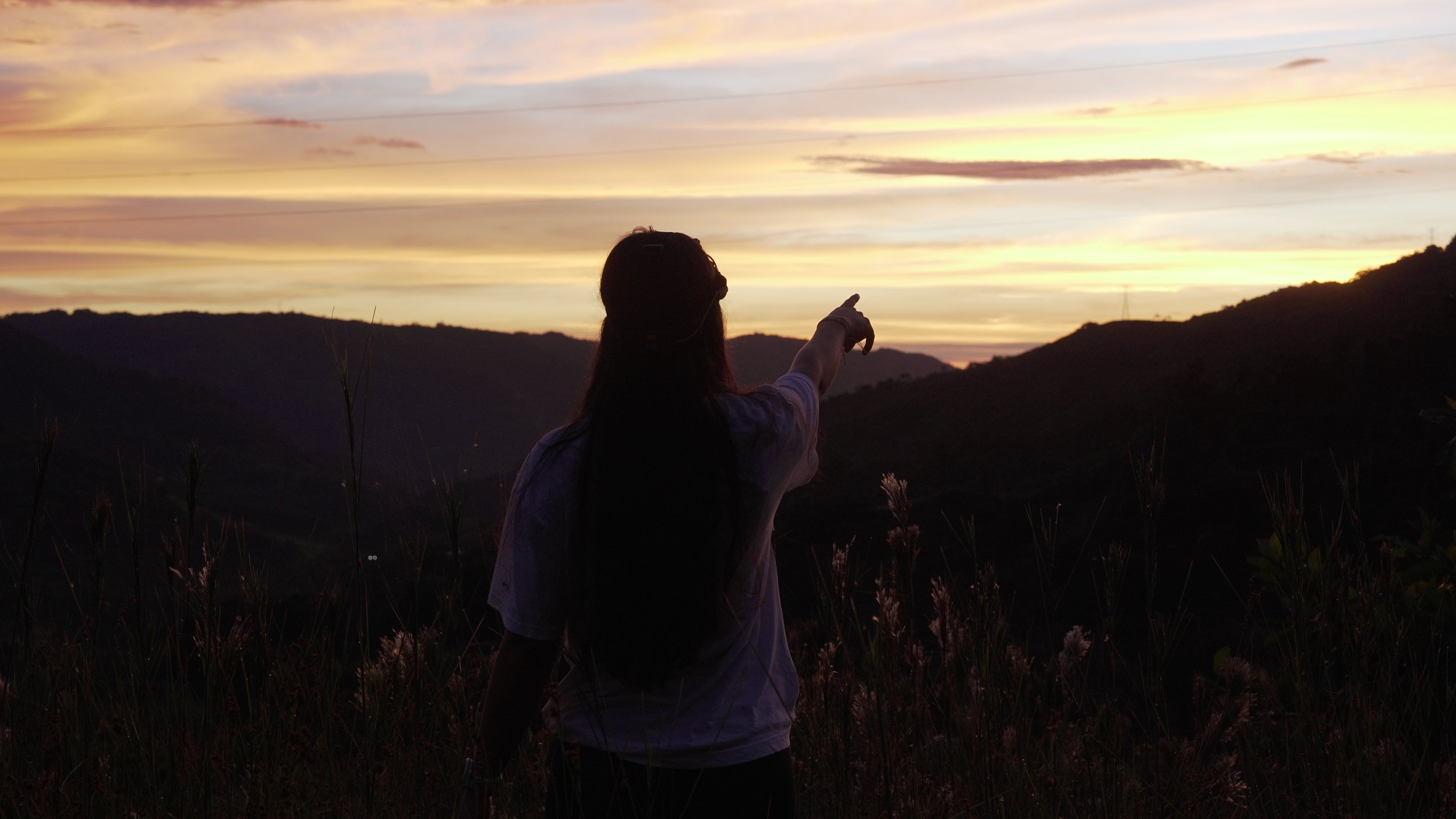 Persona apuntando hacia un atardecer en las montañas, rodeada de vegetación y nubes en tonos anaranjados y morados.