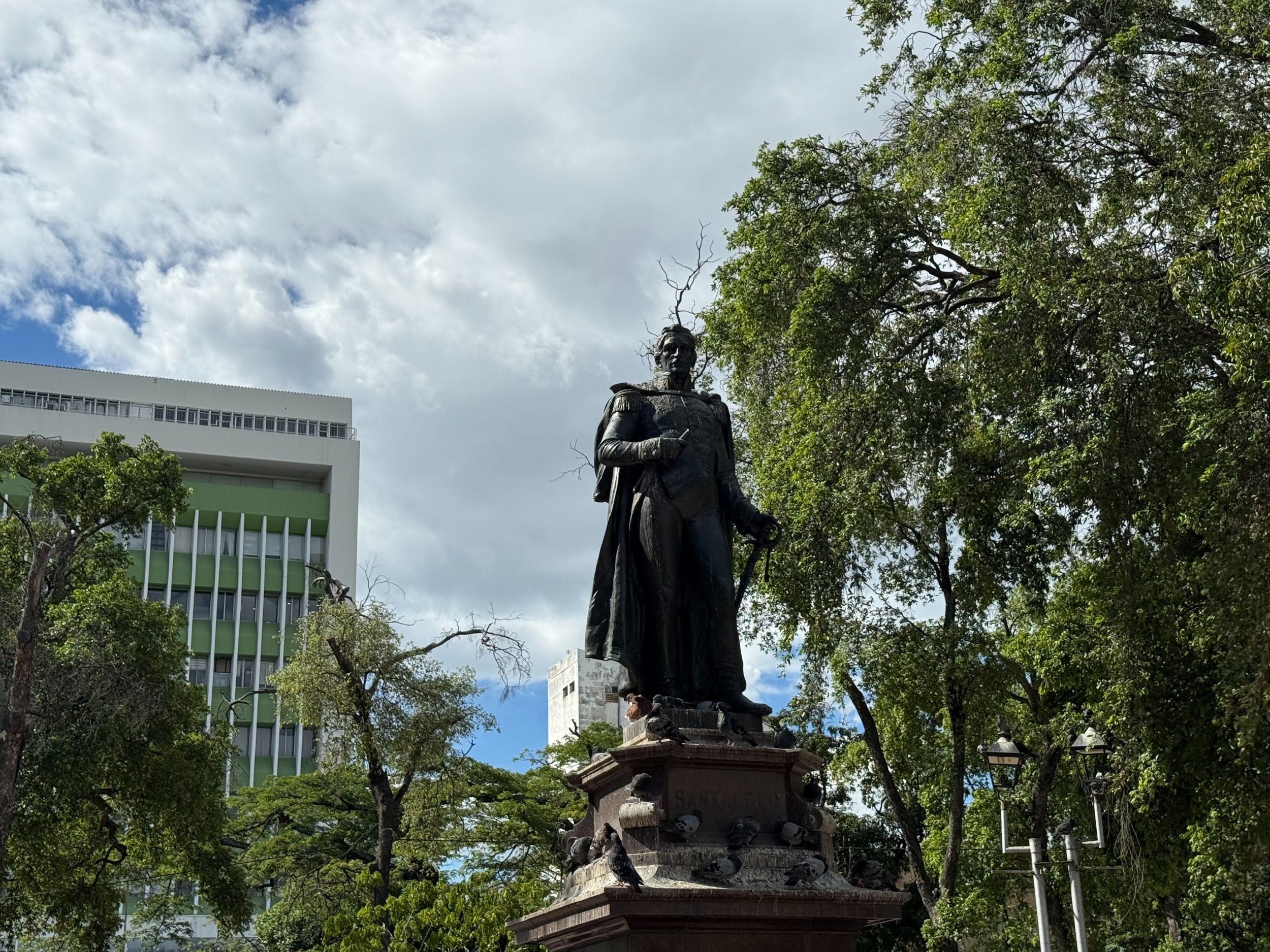Escultura de un personaje histórico en un parque, rodeada de árboles y edificios modernos al fondo, con un cielo nublado.
