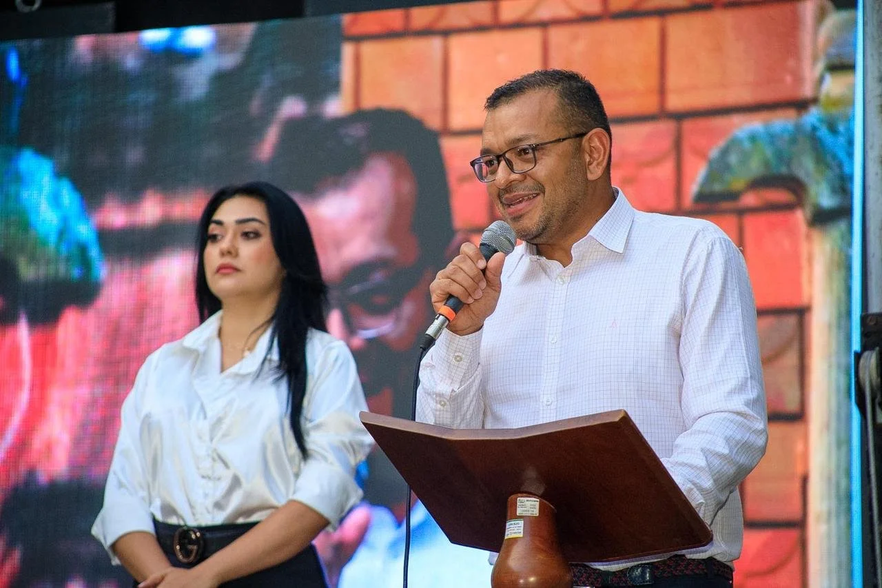 César Oliveros, un hombre con gafas y camisa blanca hablando con micrófono en un podio, acompañado por una mujer con blusa blanca y cabello oscuro, en un evento con fondo de pantalla grande y decoraciones en la pared.