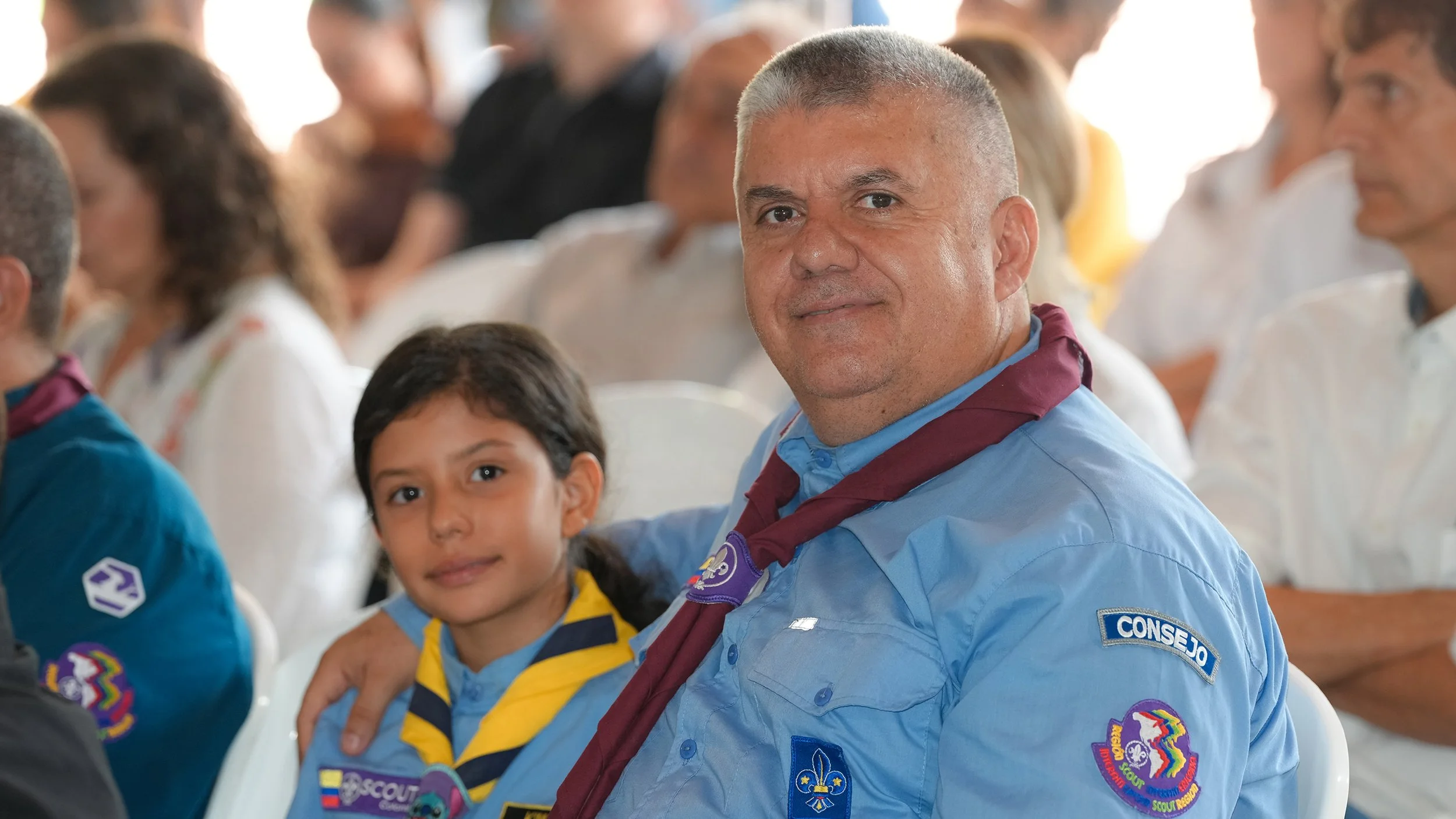 Un hombre y una niña en un evento de Scouts, ambos usan uniforme con insignias y la niña lleva un pañuelo amarillo y azul.