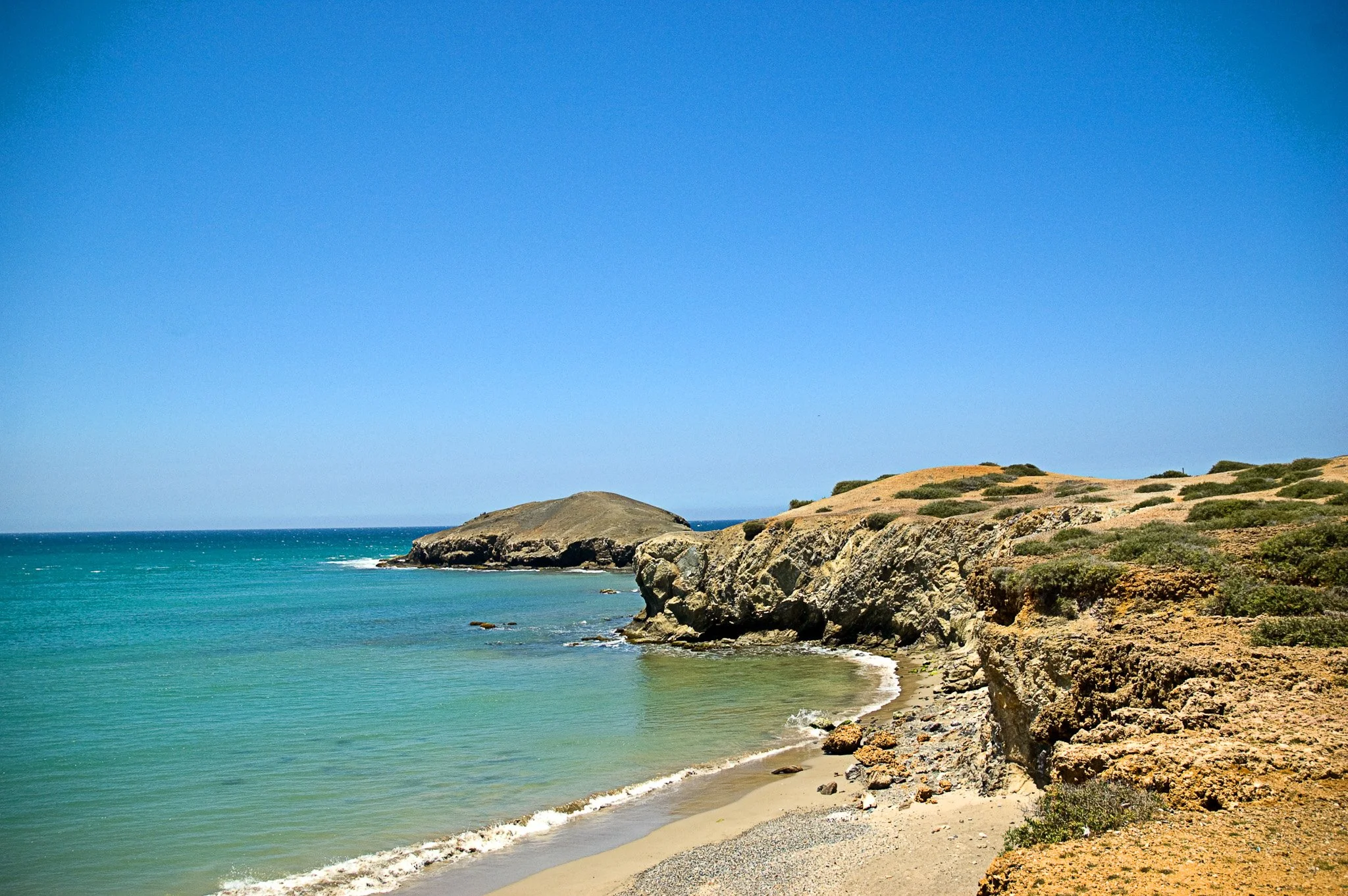 Vista de una playa con agua turquoise, rocas y colinas áridas bajo un cielo despejado.