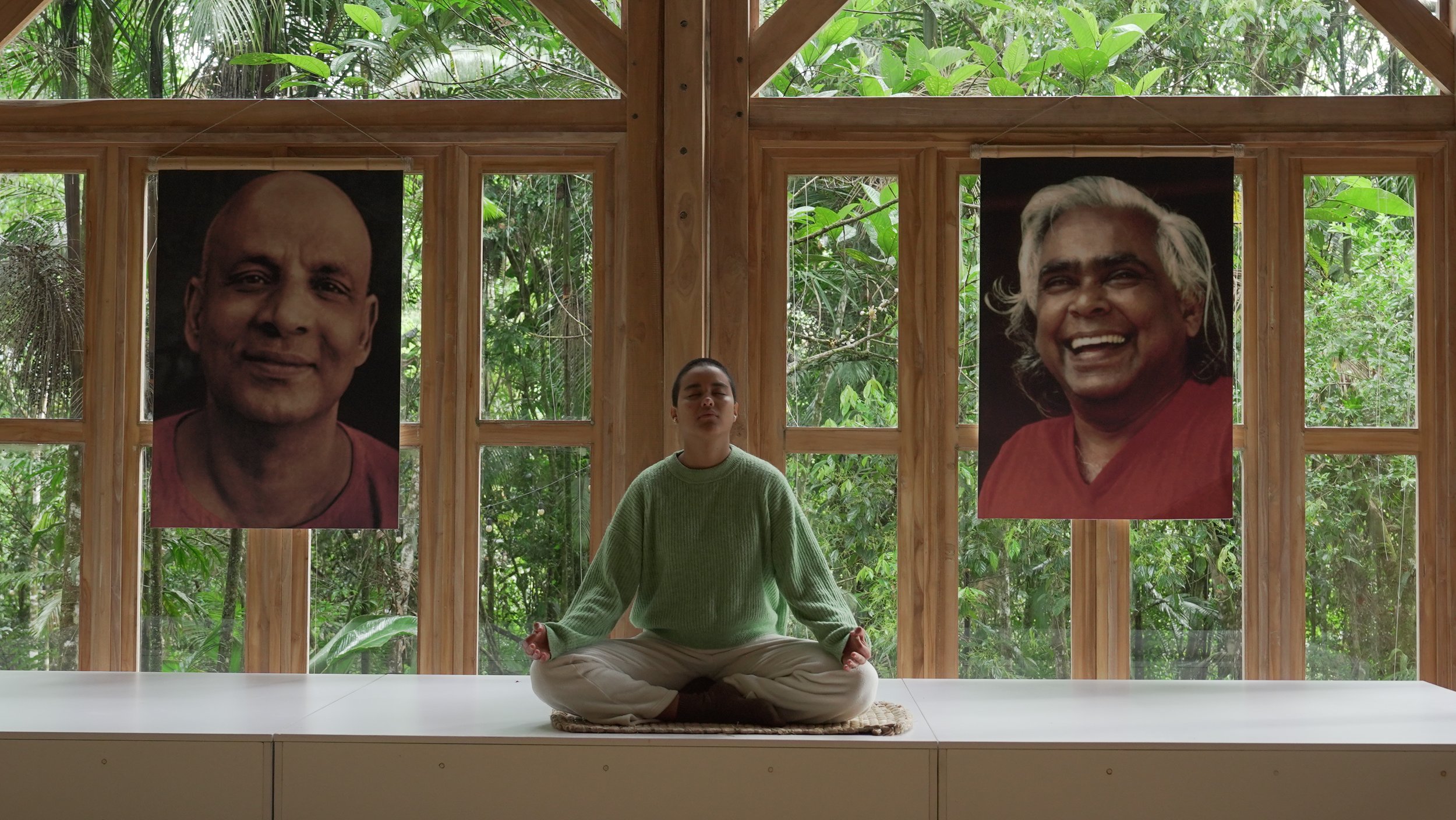 persona en posición de meditación en un espacio interior con paredes de madera y grandes ventanas, decoradas con retratos de personas sonrientes, rodeada de vegetación exterior