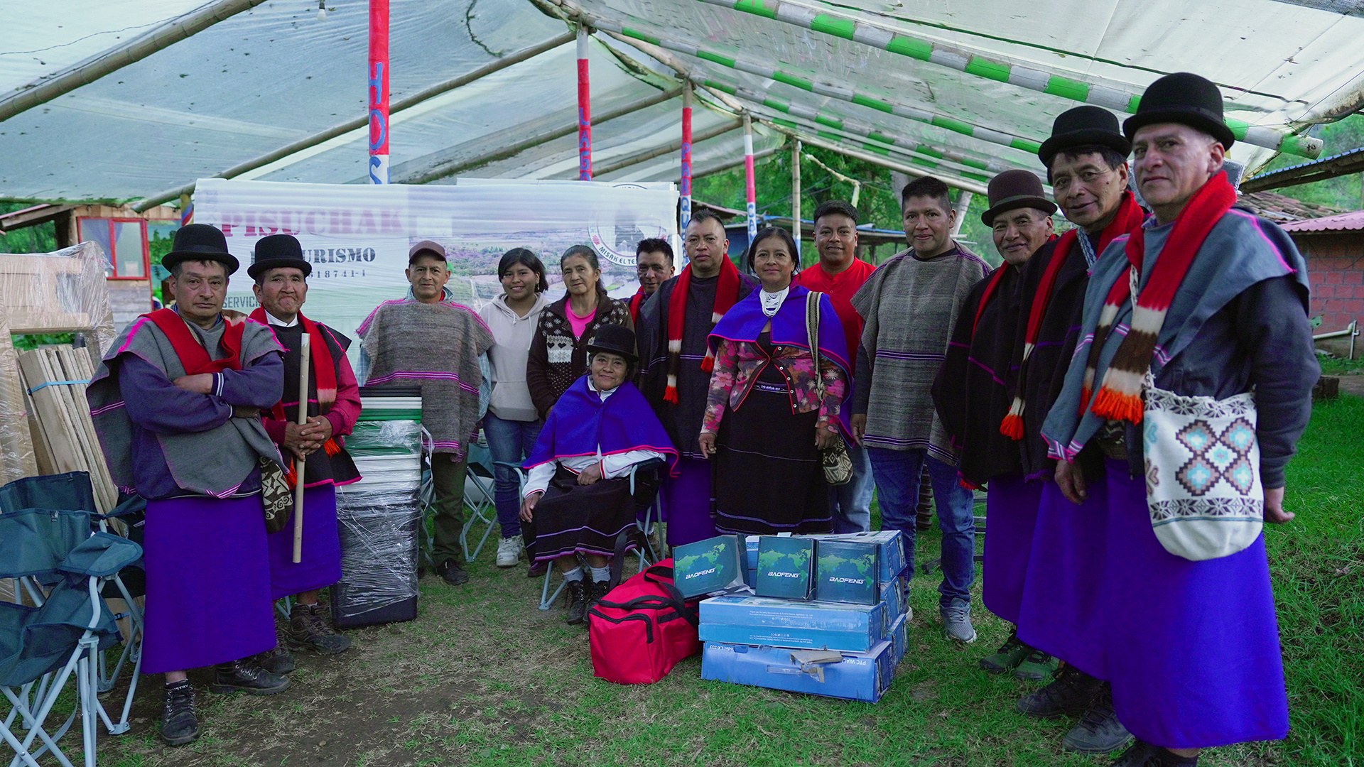 Grupo de personas vestidas con ropa tradicional indígena en un ambiente al aire libre, con objetos y regalos en el suelo.