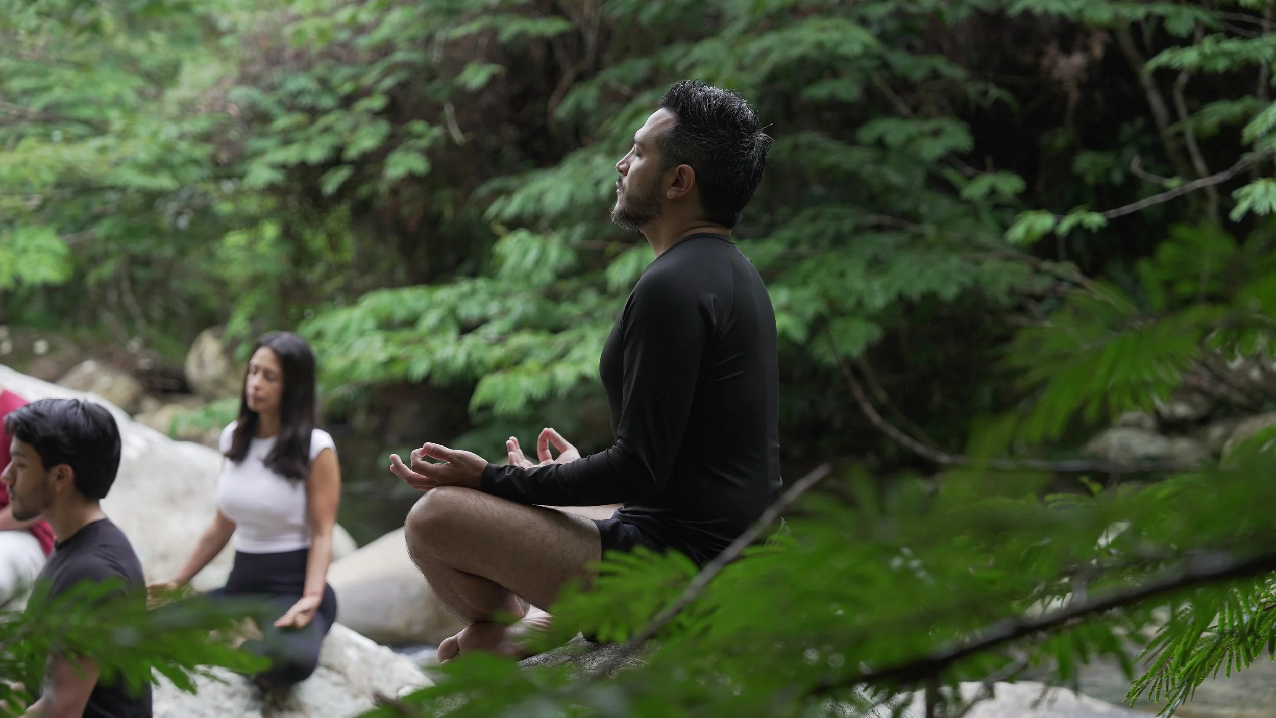 Un hombre realiza una meditación en postura de loto en un entorno natural junto a un río, rodeado de vegetación verde, mientras otras personas también meditan en el fondo.