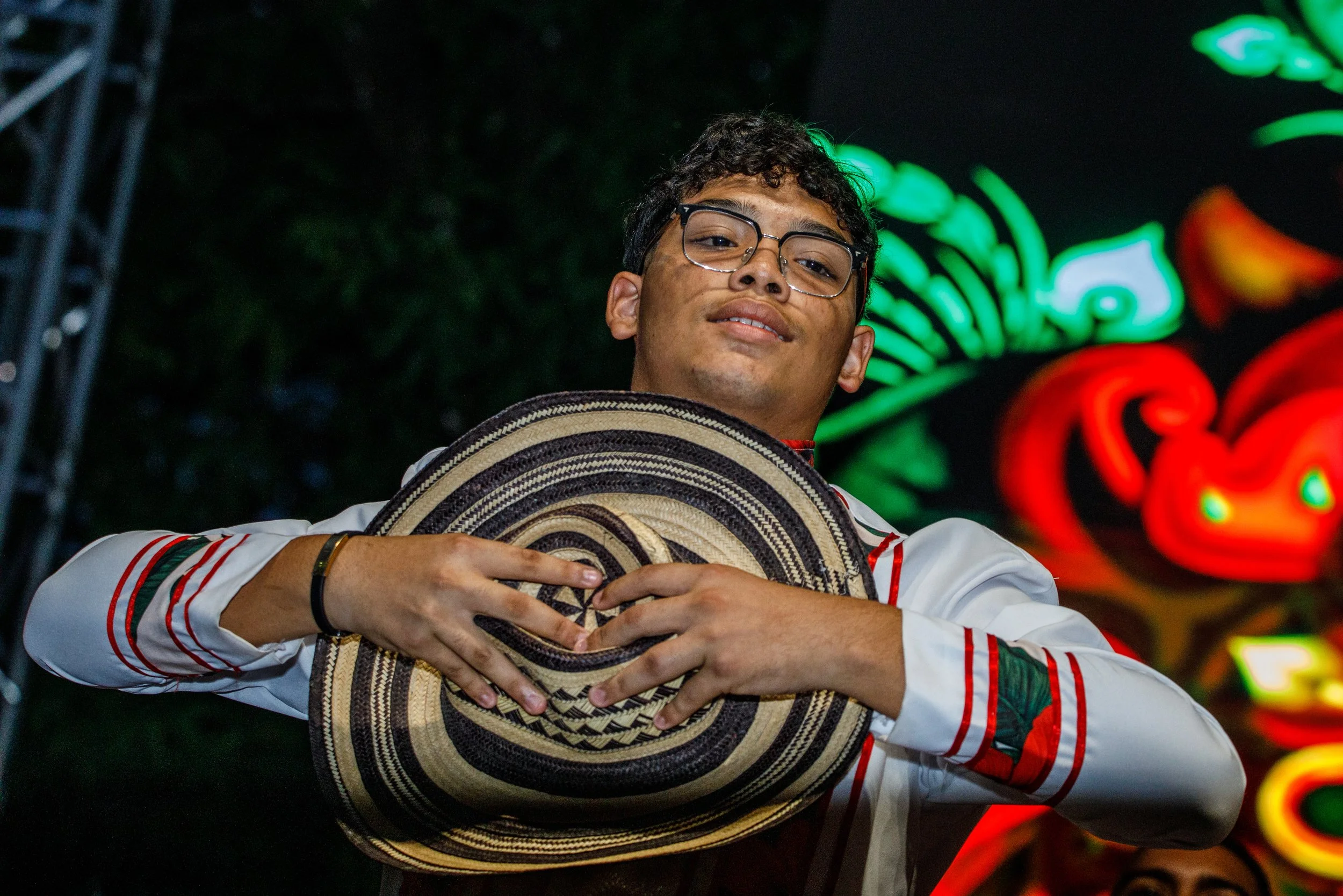 Joven con gafas sosteniendo un sombrero de usulután frente a fondo de luces de neón en la noche