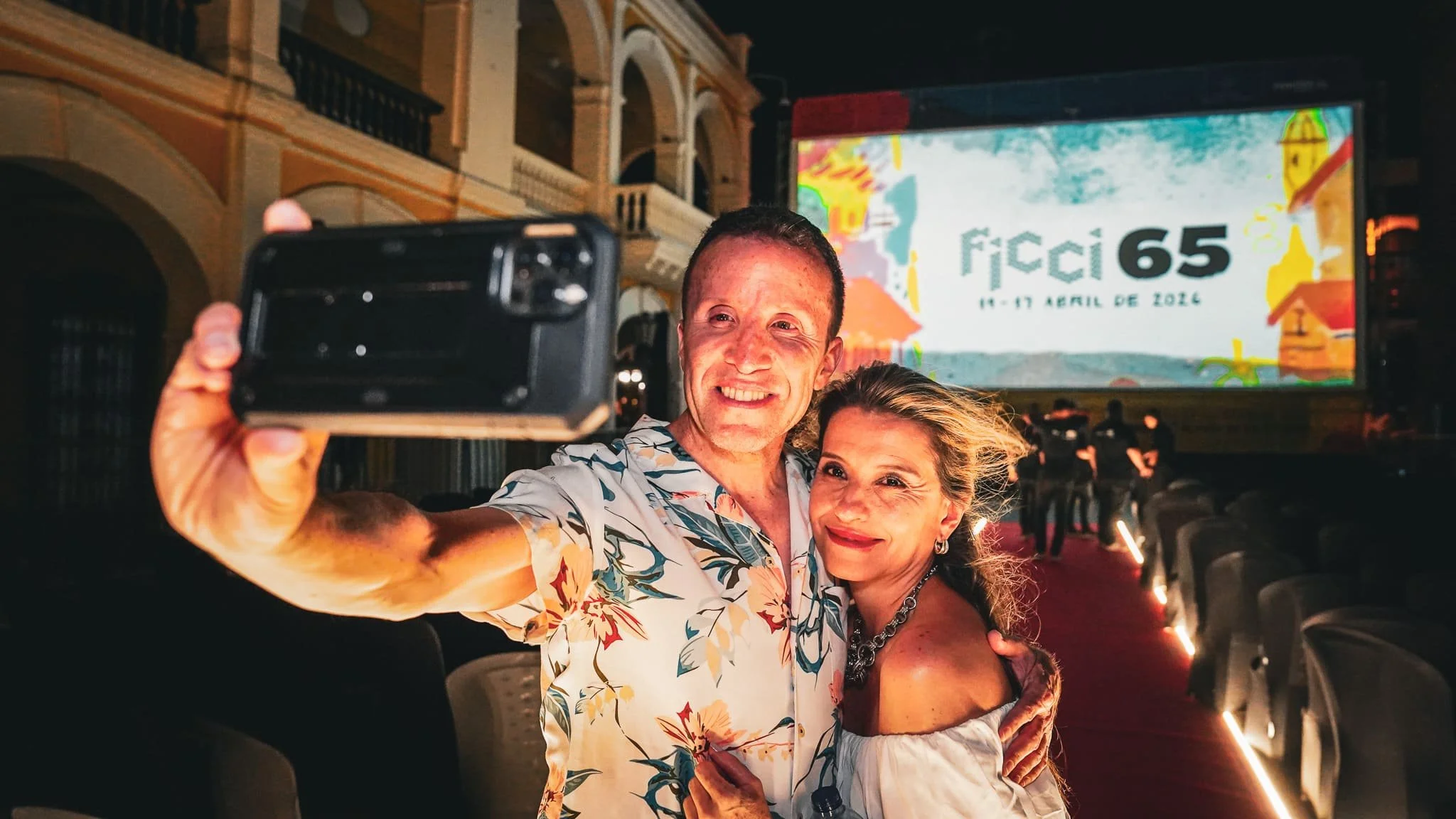 Una pareja sonriendo toma una selfie en un evento llamado FICCI 65, que se celebra del 11 al 17 de abril de 2026, en un lugar con arquitectura colonial. Hay sillas y una pantalla con el logo del evento.