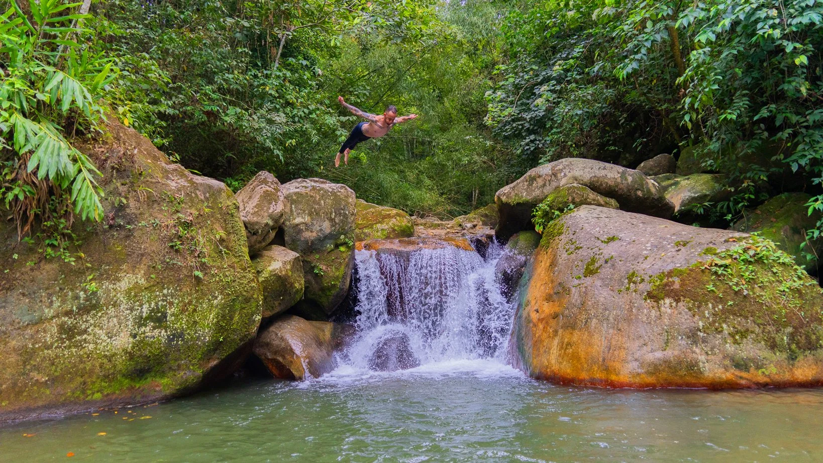 Una persona saltando sobre una cascada en un río rodeado de vegetación densa y rocas grandes.