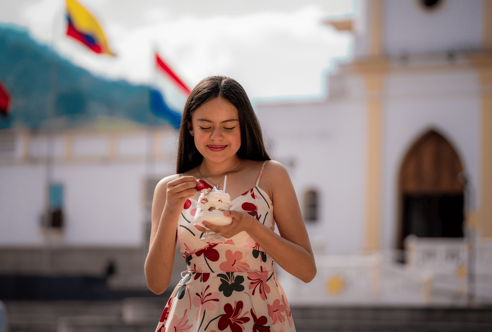 Una joven con vestido de verano con estampado floral sostiene un helado en un lugar público. Detrás, banderas flotan en el aire y edificios blancos adornados con detalles en colores claros, en un día nublado.