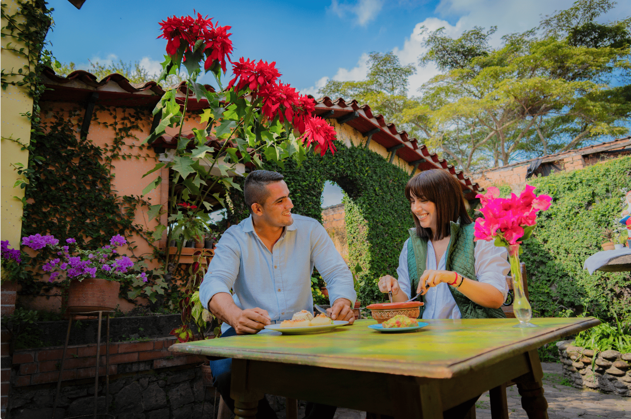 Hombres y mujer sonriendo en una mesa de desayuno en un jardín con muchas plantas y flores, con árboles y cielo despejado en el fondo.