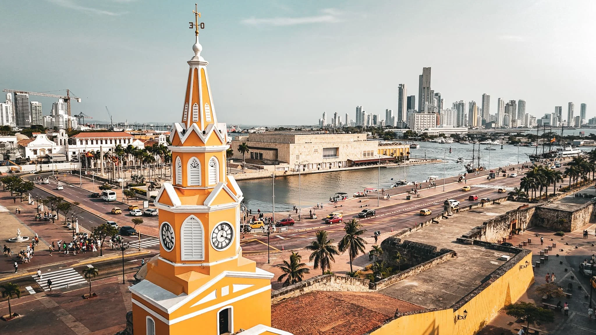Vista de la ciudad de Cartagena, Colombia, con la torre del reloj amarilla en primer plano, el mar y los rascacielos en el fondo, y un muelle con barcos y personas caminando a lo largo del paseo marítimo.