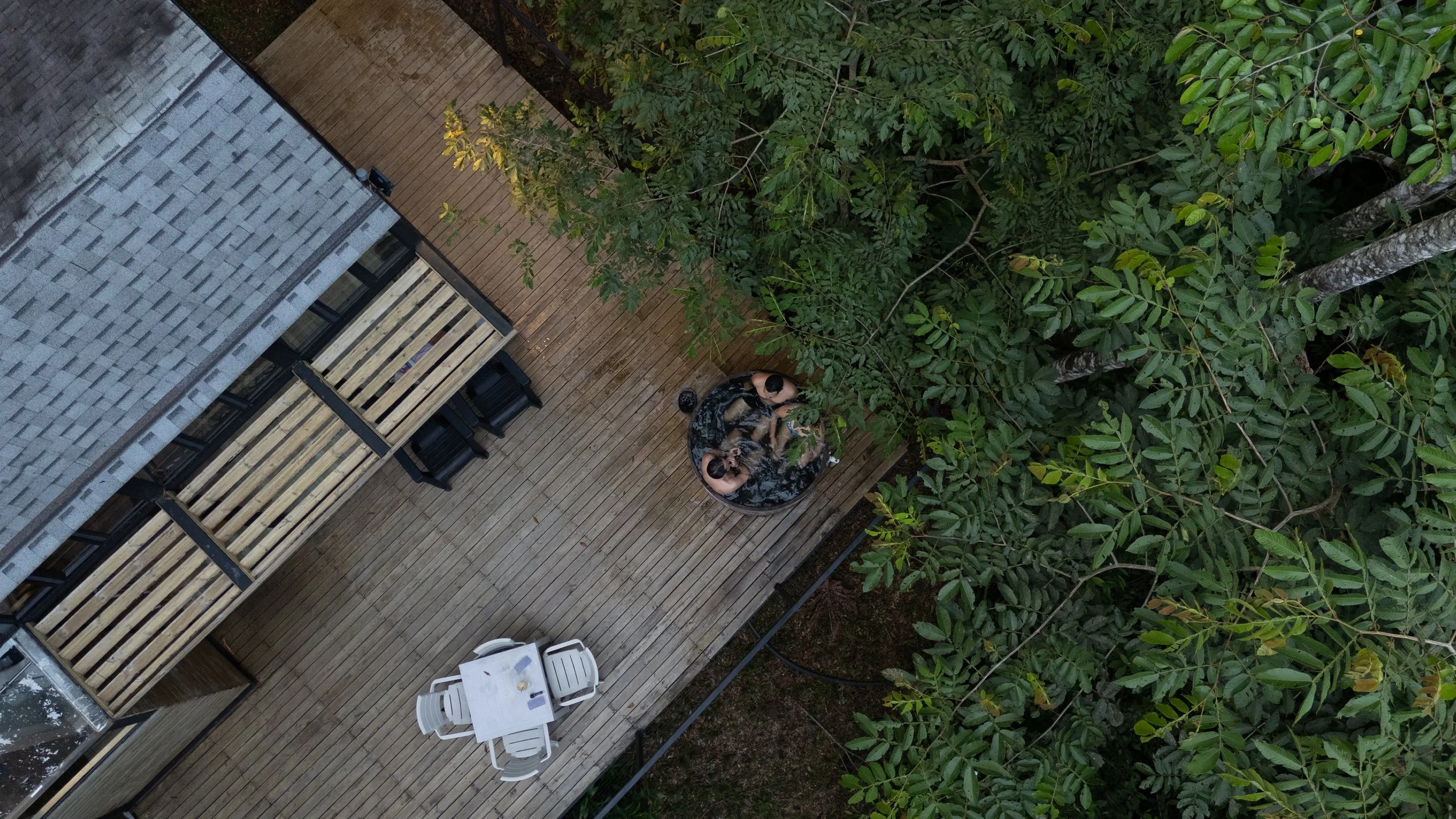 Vista aérea de una terraza de madera con una mesa redonda, sillas blancas, y una familia de tres personas sentadas hablando, rodeada de árboles verdes.
