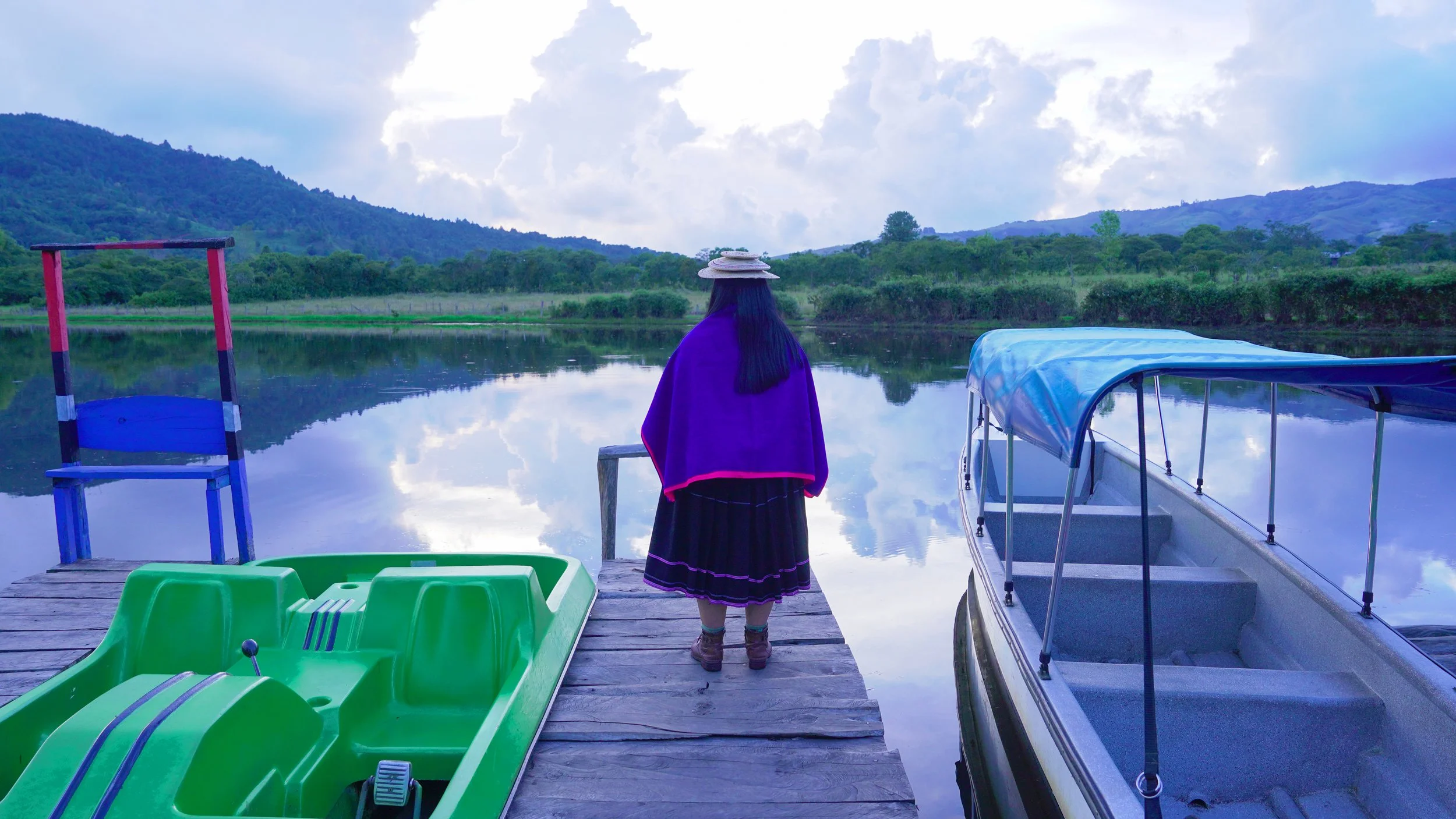 Una mujer con sombrero y ropa tradicional de colores oscuros de espaldas, mira un lago en una zona rural rodeada de montañas y árboles, con barcos y juegos infantiles en la escena.