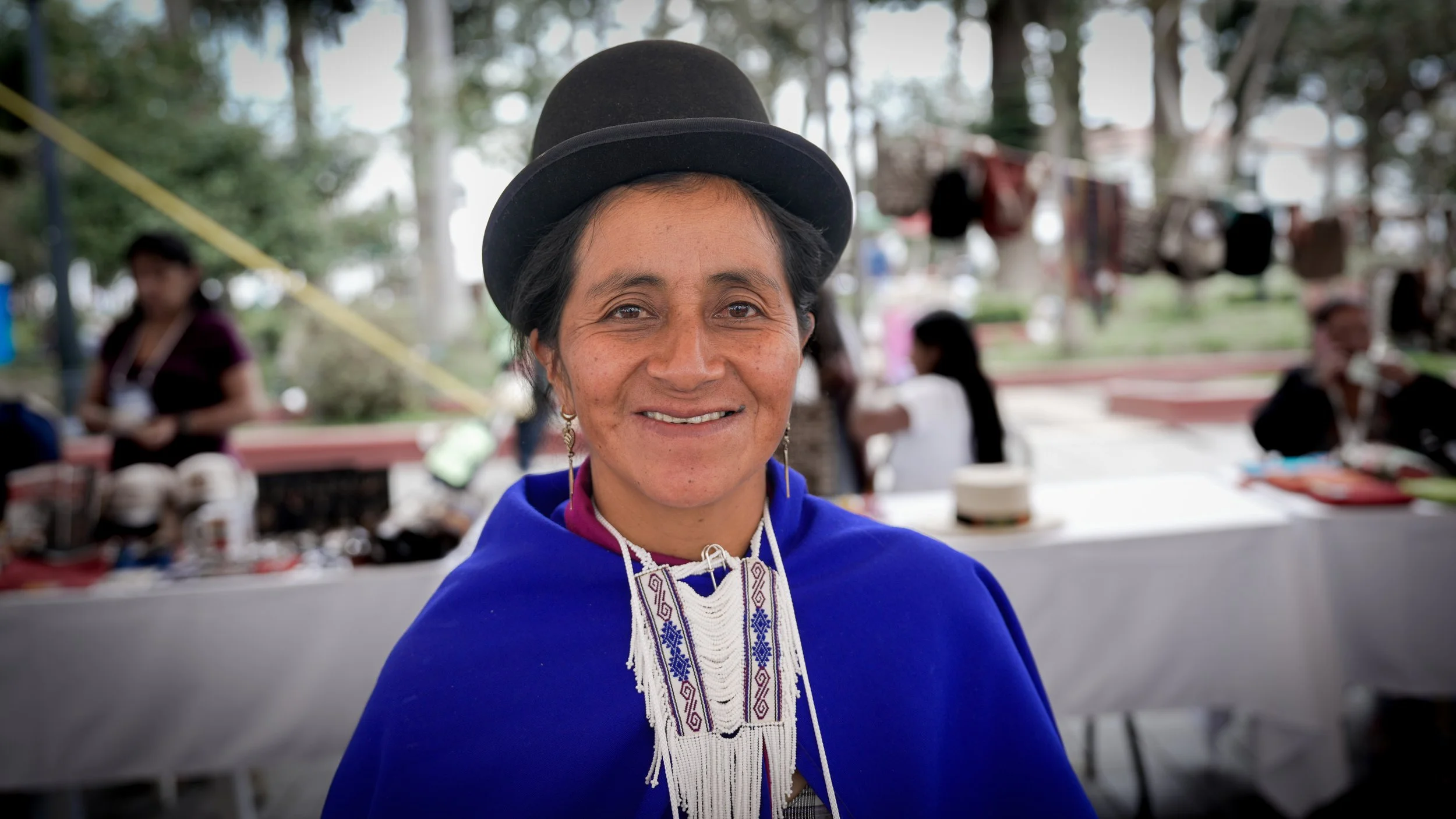 Mujer indígena sonriendo, lleva un sombrero negro y una blusa azul, en un ambiente al aire libre con varias personas y árboles de fondo.