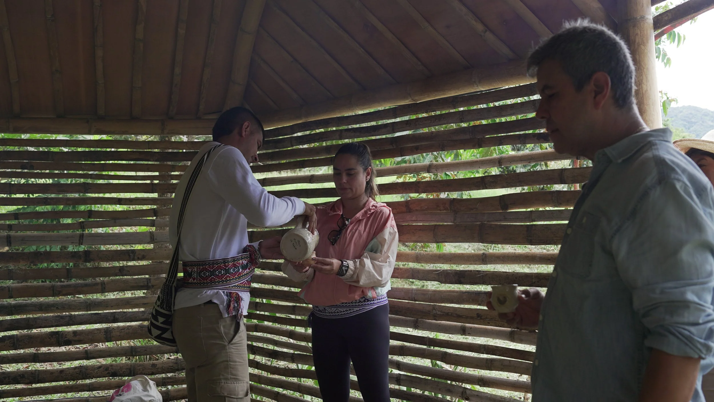 Cuatro personas en un espacio rústico de madera, una mujer recibe un objeto de un hombre con un diseño tradicional, mientras otros observan, en un entorno natural.