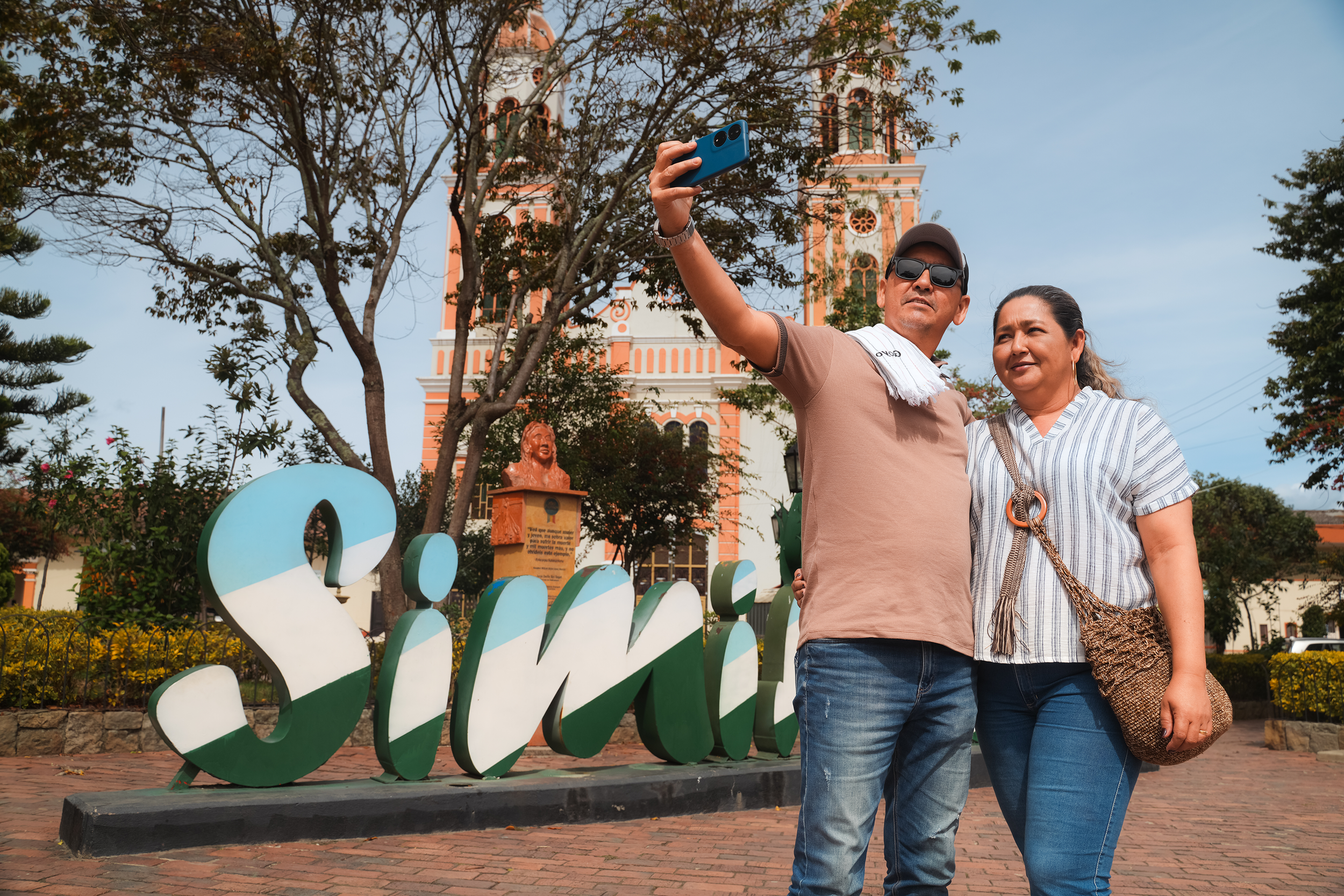 Una pareja toma una selfie frente a un edificio color coral con campanarios y ventanas arqueadas, rodeados de árboles y plantas en un parque urbano.