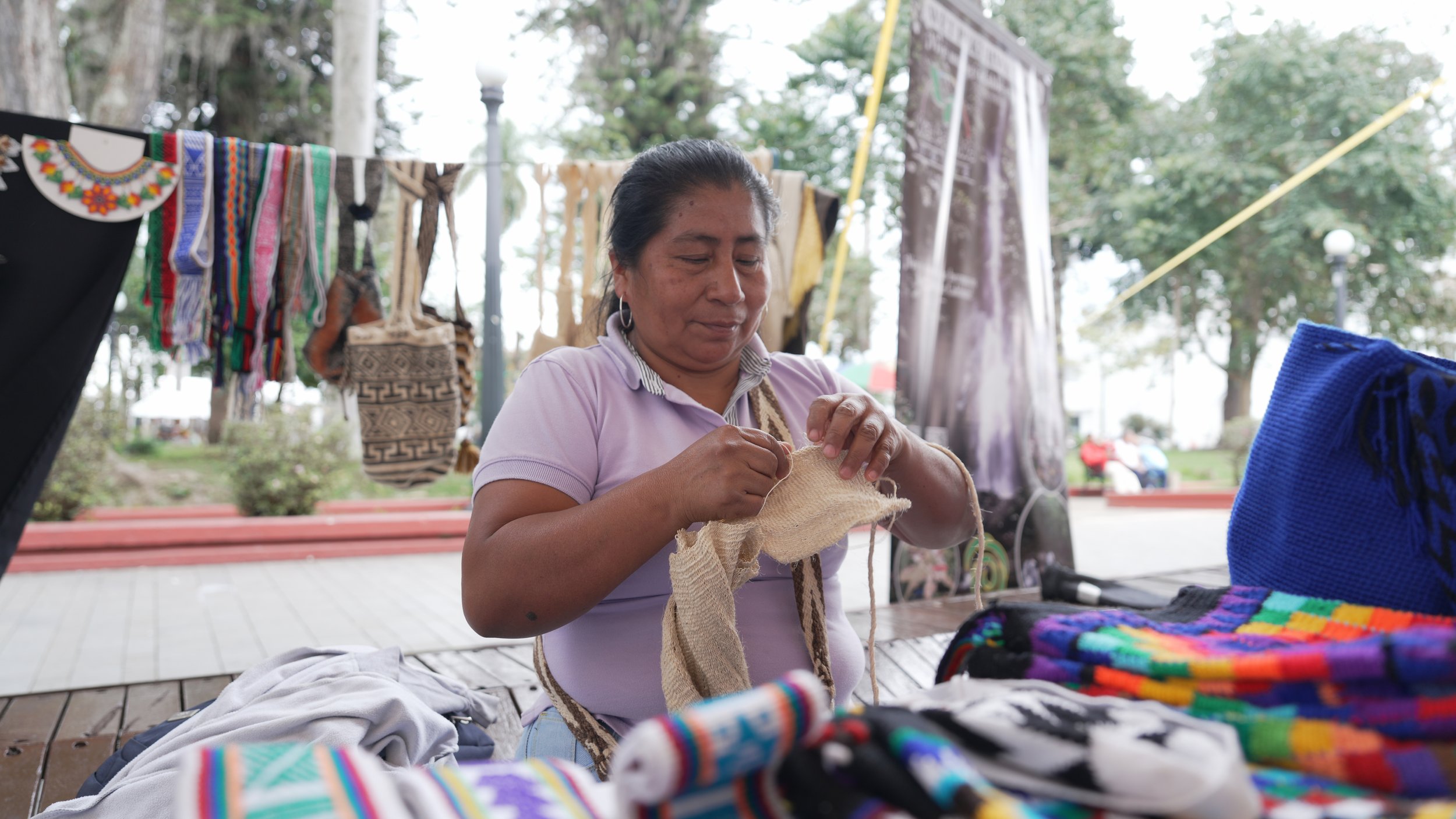 Mujer indígena tejiendo con fibra de color natural en un mercado al aire libre con telas y prendas coloridas a su alrededor.