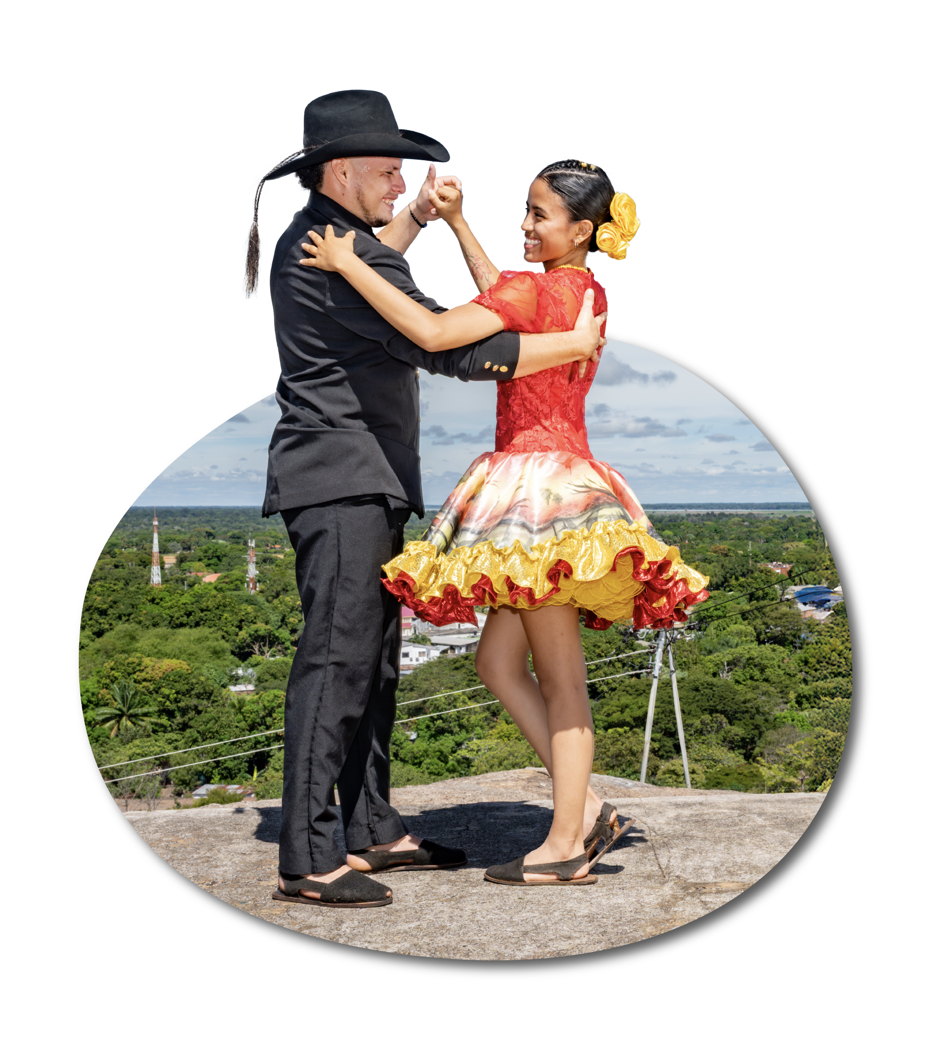 Pareja bailando junte en una loma con vista a campo y casas, ella con vestido colorado y falda floreada, él con sombrero y traje oscuro, ambos sonriendo y enroscados en un abrazo.