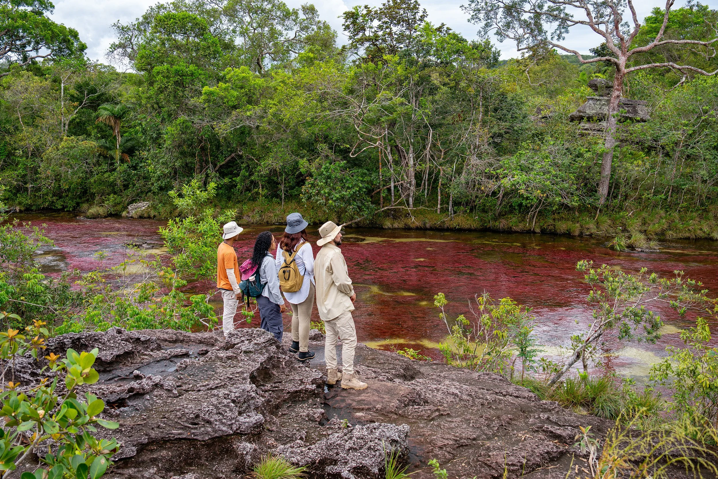 Grupo de cinco personas con sombreros explorando un bosque tropical cerca de un río con agua de color rojo.