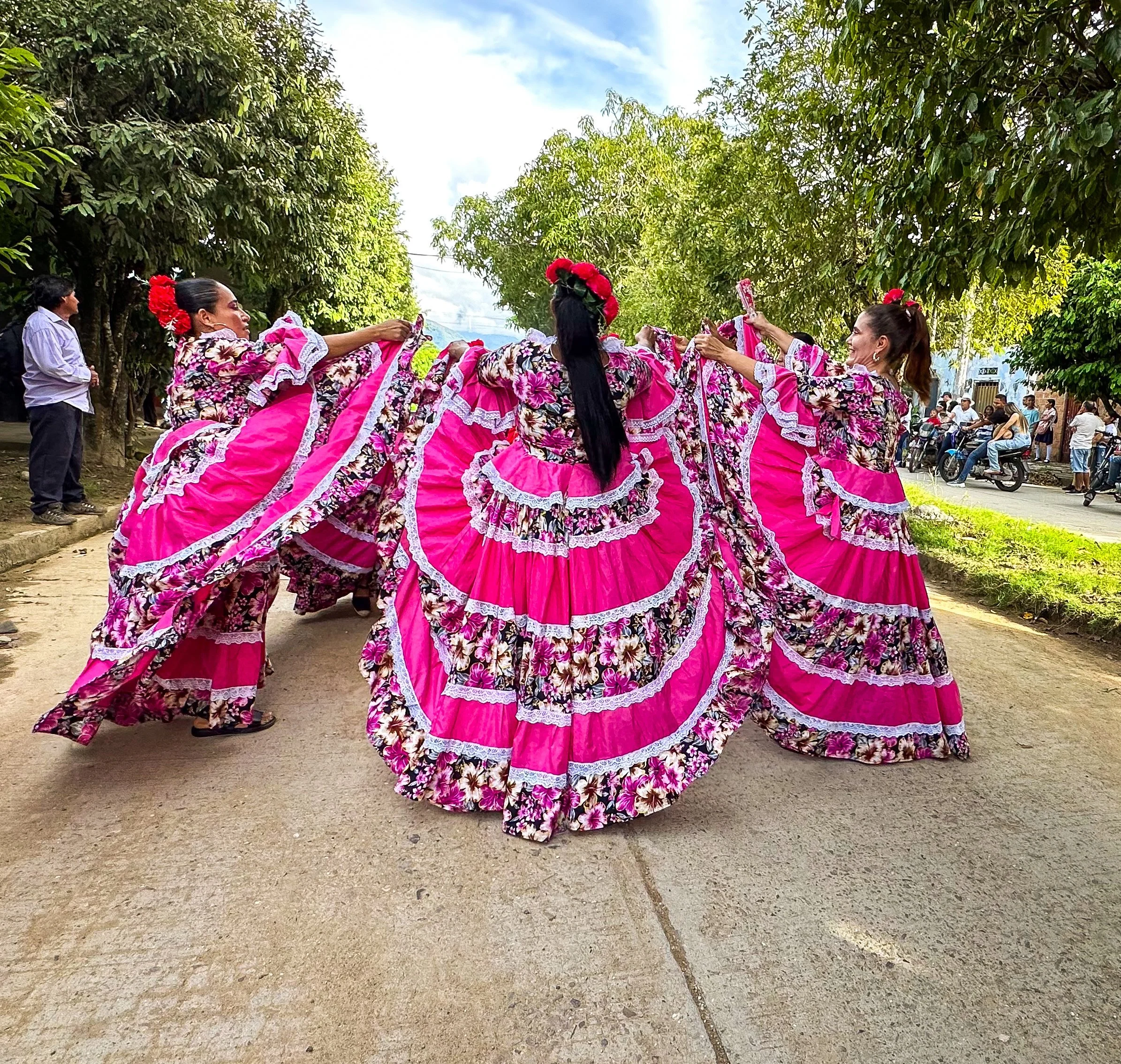 Grupo de mujeres bailando en una fiesta tradicional mexicana, usando vestidos color rosa con flores y encajes, en un ambiente al aire libre con árboles y personas observando.