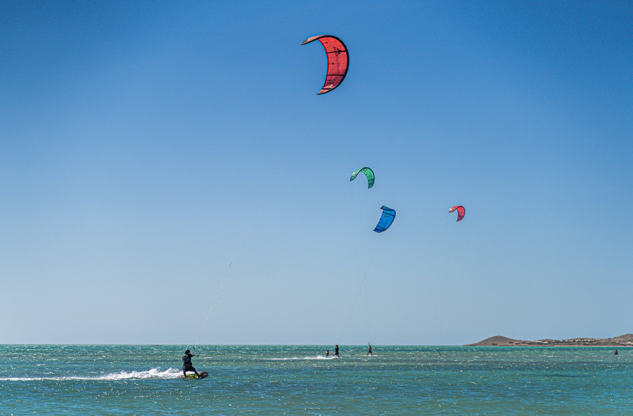 Cuatro personas practicando kitesurf en el mar con cometas de colores en el cielo azul y un paisaje costero en el fondo.