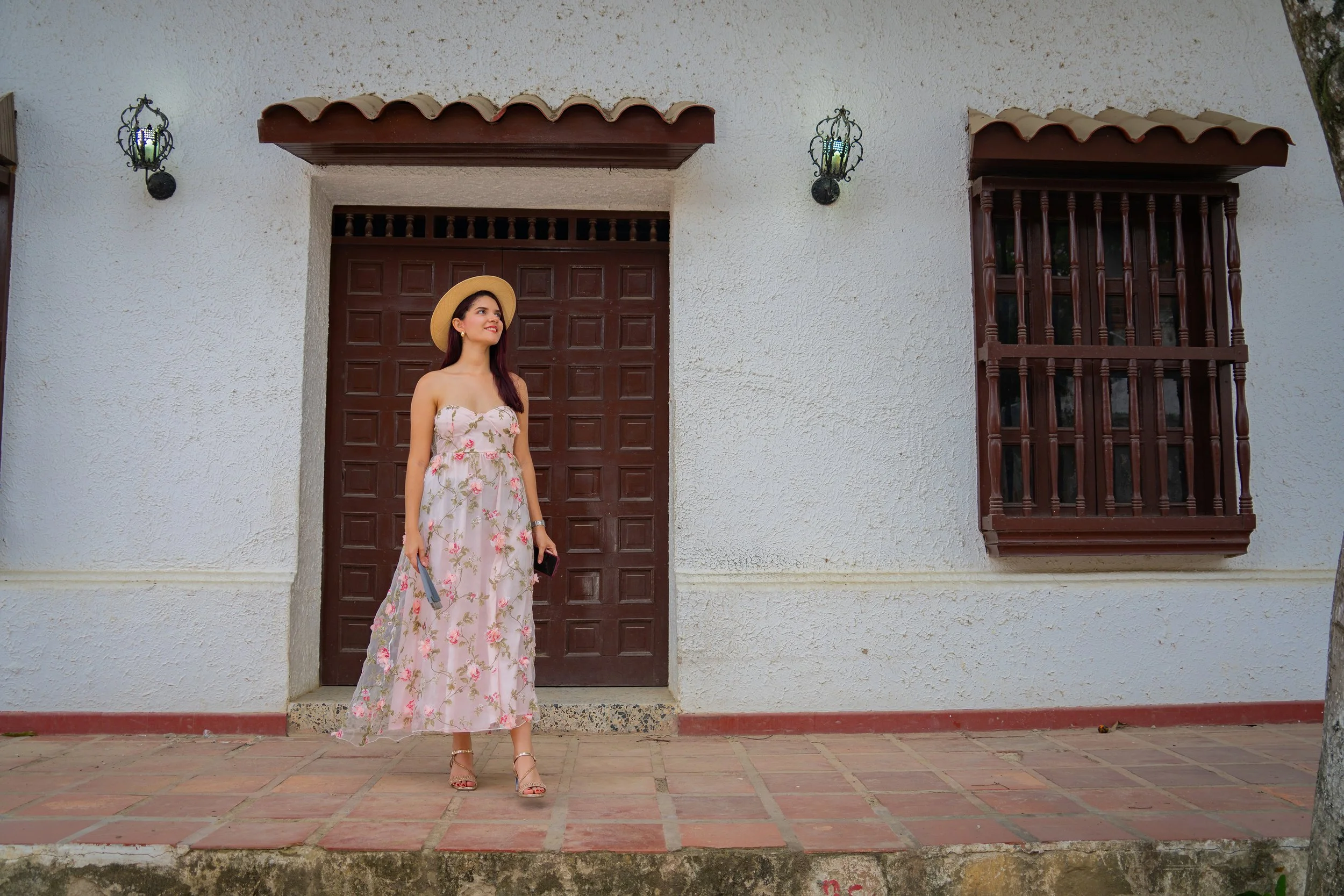 Mujer con vestido largo de flores y sombrero, de pie frente a una puerta de madera en una casa blanca.