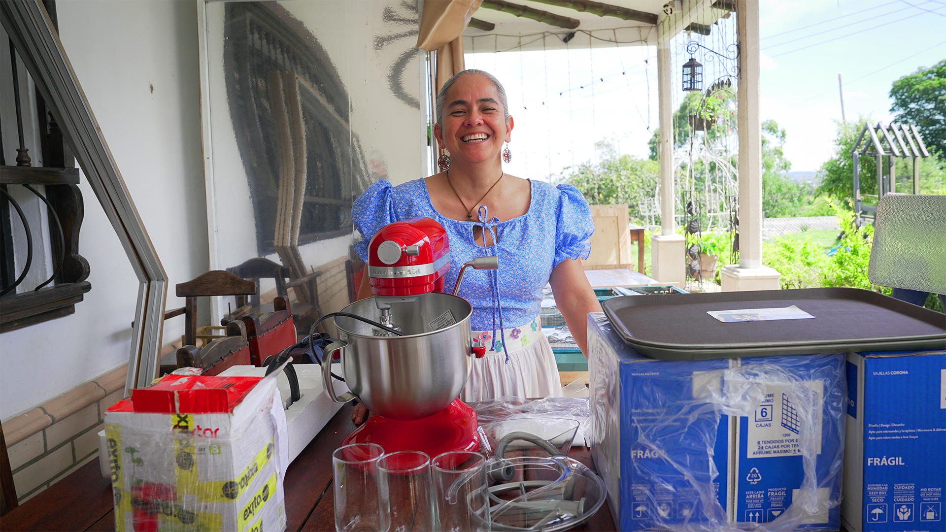 Mujer sonriente con camiseta azul y falda clara en una terraza rodeada de plantas, con electrodomésticos de cocina y cajas almacenadas en una mesa, en un día soleado.