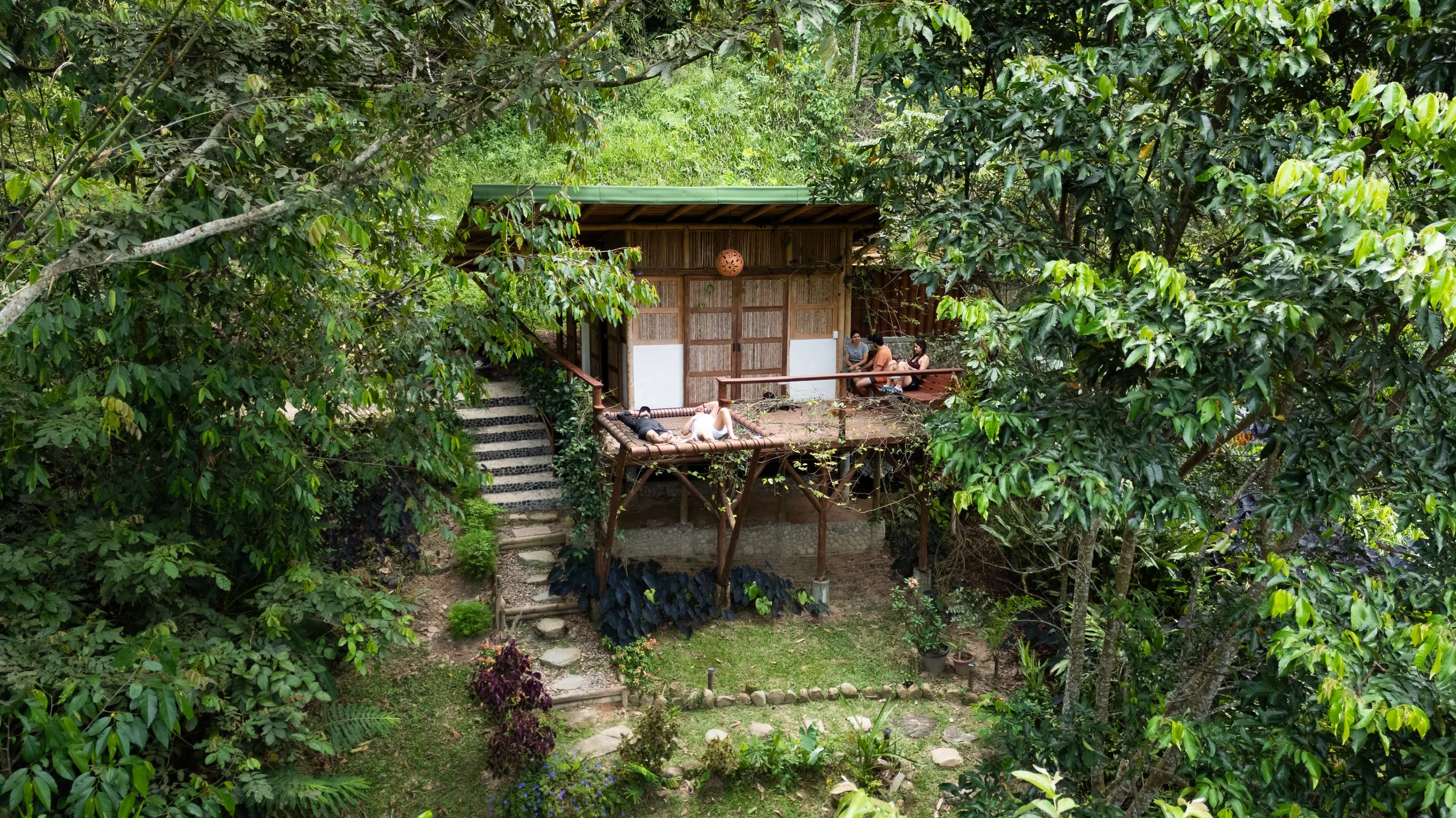 Casa de madera en medio de un bosque con muchas plantas y árboles, personas descansando en el balcón y en una hamaca.