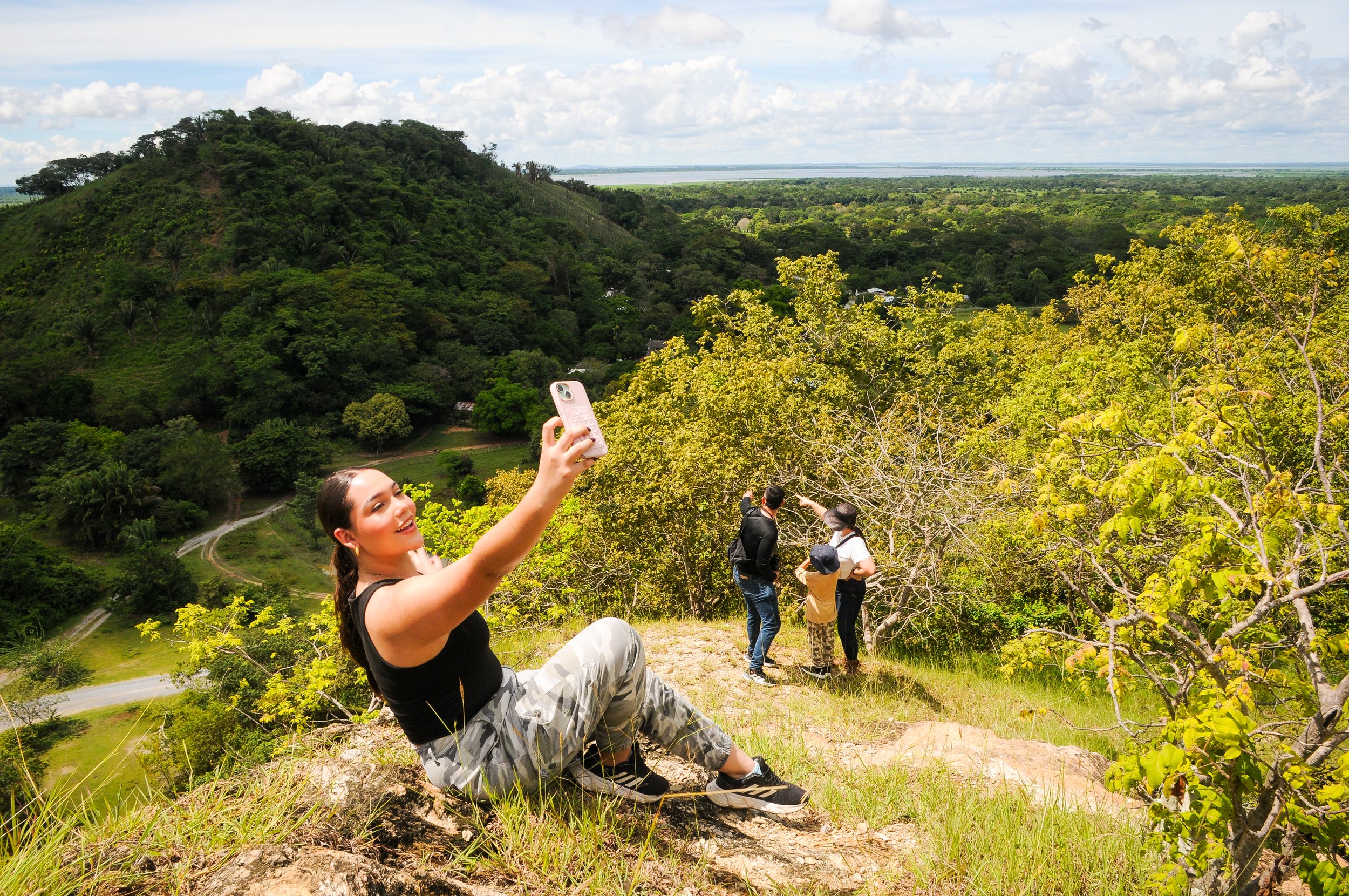 Grupo de turistas en una colina tomando fotos con teléfonos móviles, rodeados de vegetación lush y vista panorámica de un paisaje selvático y ríos lejanos.