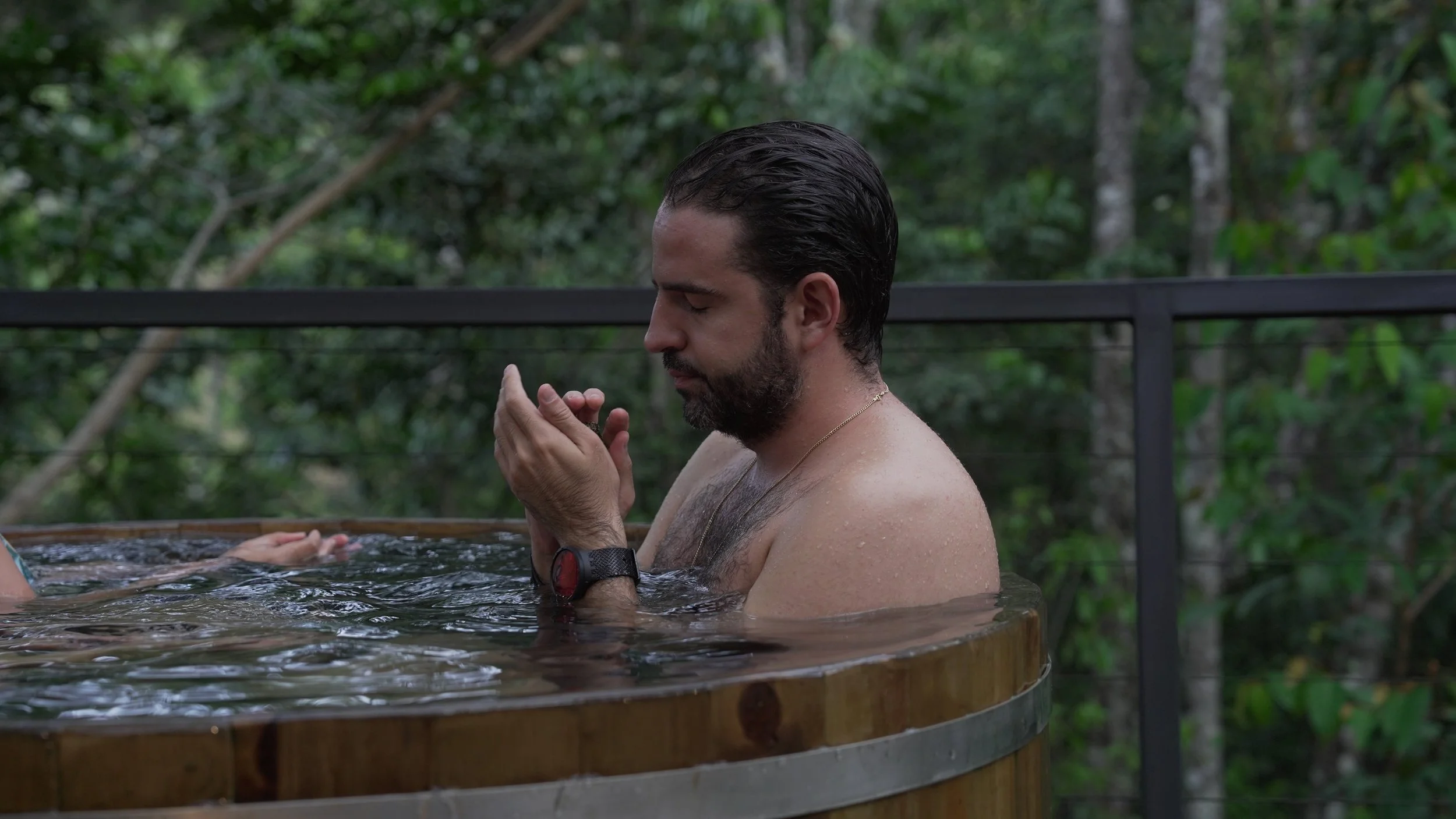 Hombre con barba y cabello oscuro en una tina de agua, con las manos juntas en señal de oración, en un entorno natural con árboles y una barandilla metallicá.