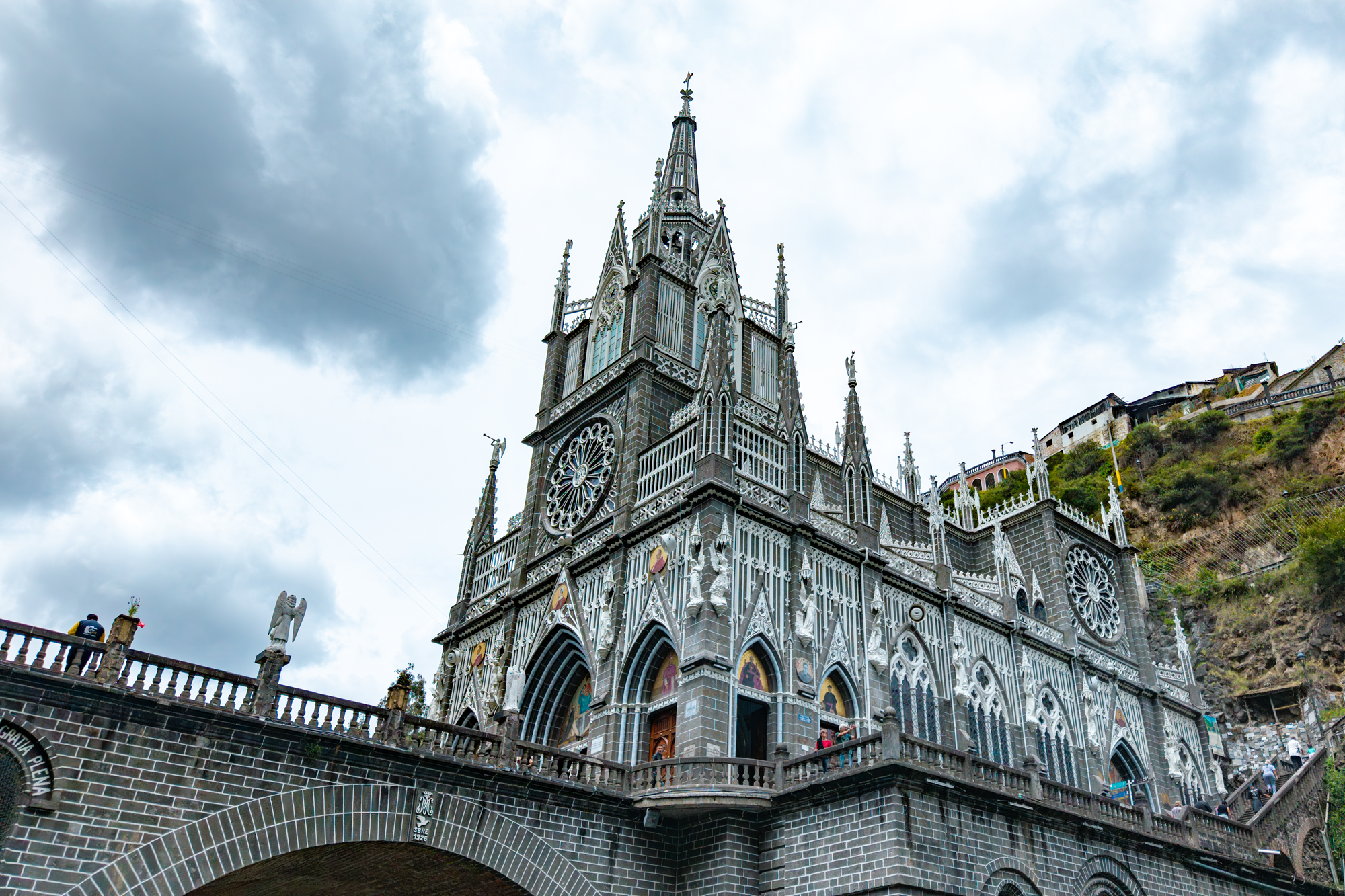 Vista de una iglesia gótica de color gris oscuro con muchos detalles arquitectónicos, situada en una colina con casas y árboles alrededor, bajo un cielo nublado.