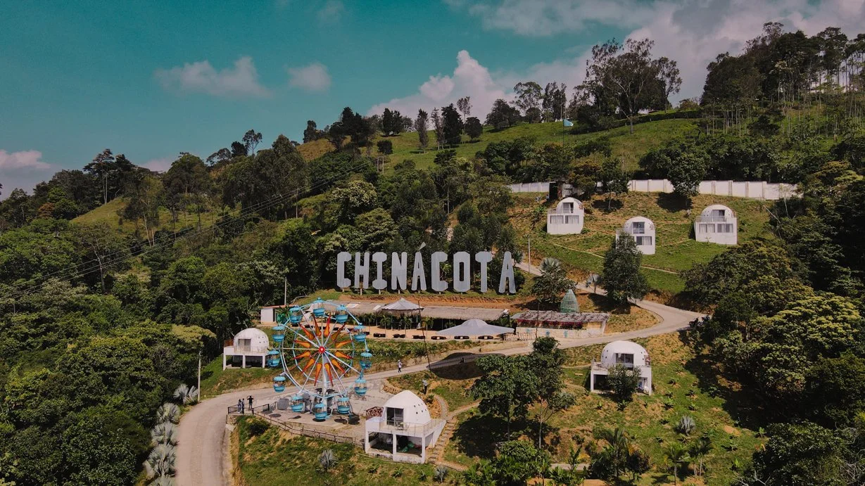 Vista aérea de Chinácota en un paisaje montañoso con casas blancas en varias elevaciones, un carrusel y un cartel grande con la palabra 'CHINACOTA' en la ladera de la colina.