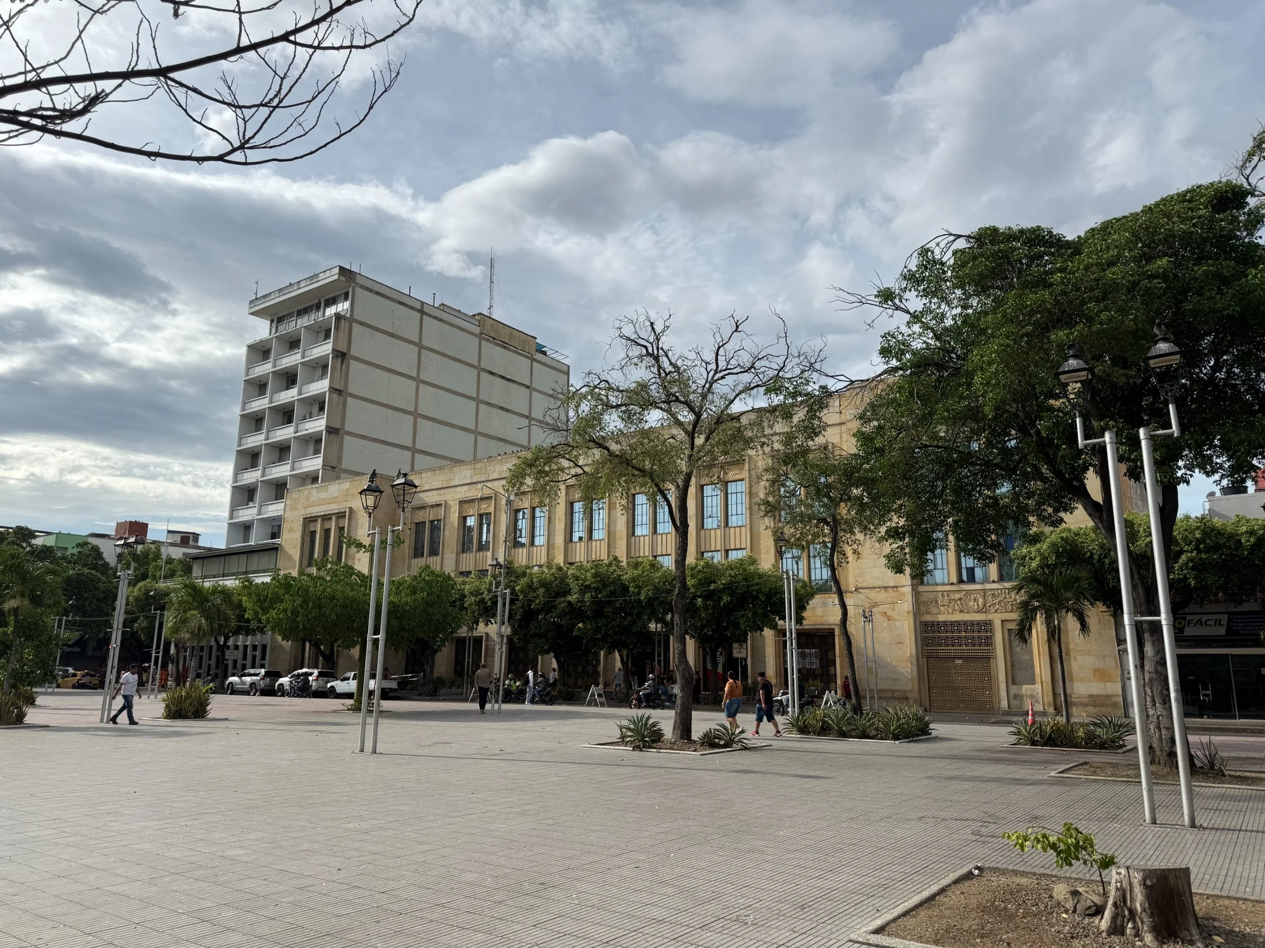 Una plaza en una ciudad con árboles, postes de luz y un edificio alto de color claro. Varias personas caminan y hay motocicletas estacionadas.
